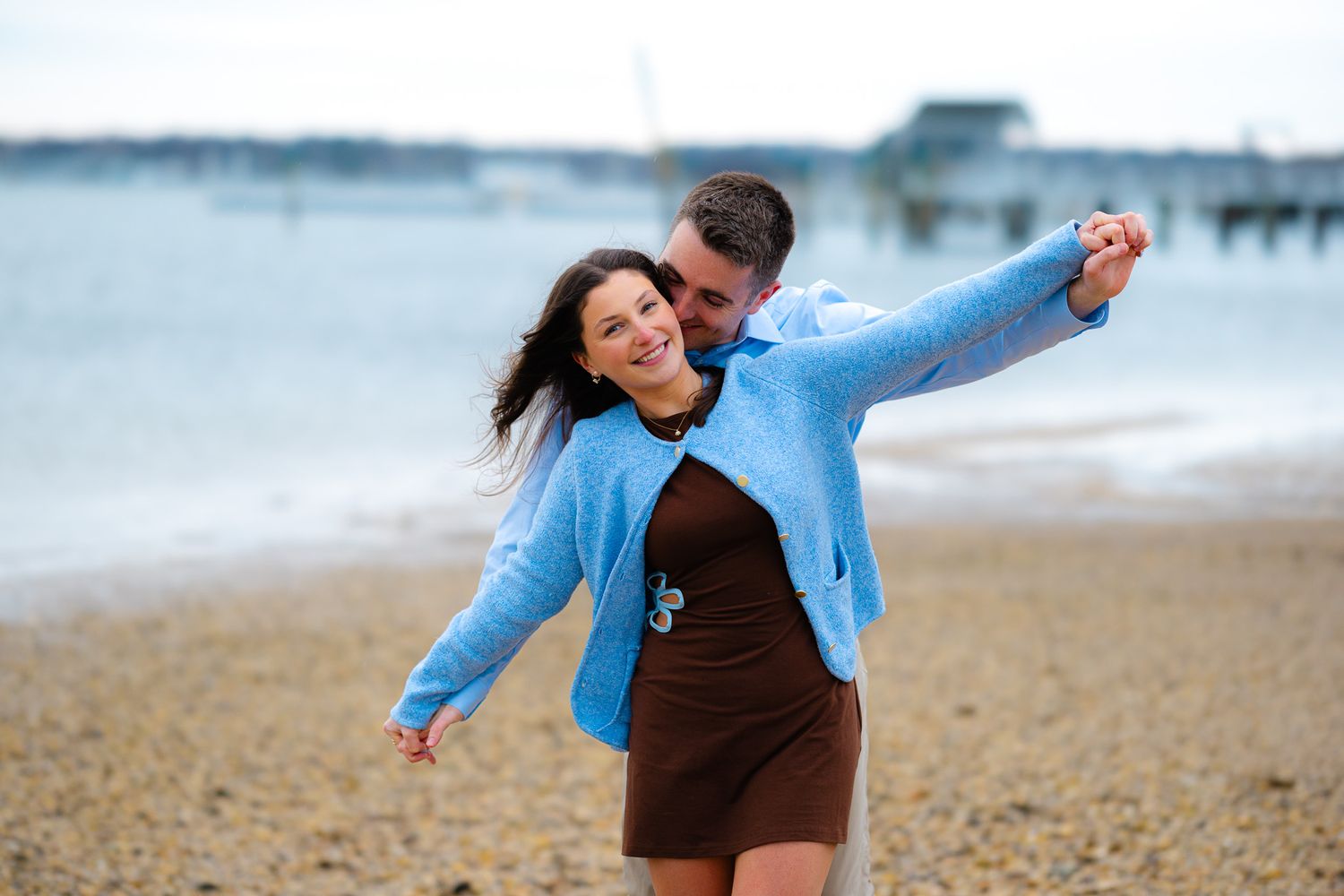 Happy couple embracing on a sandy beach, woman in brown dress and blue cardigan, pier visible in background.