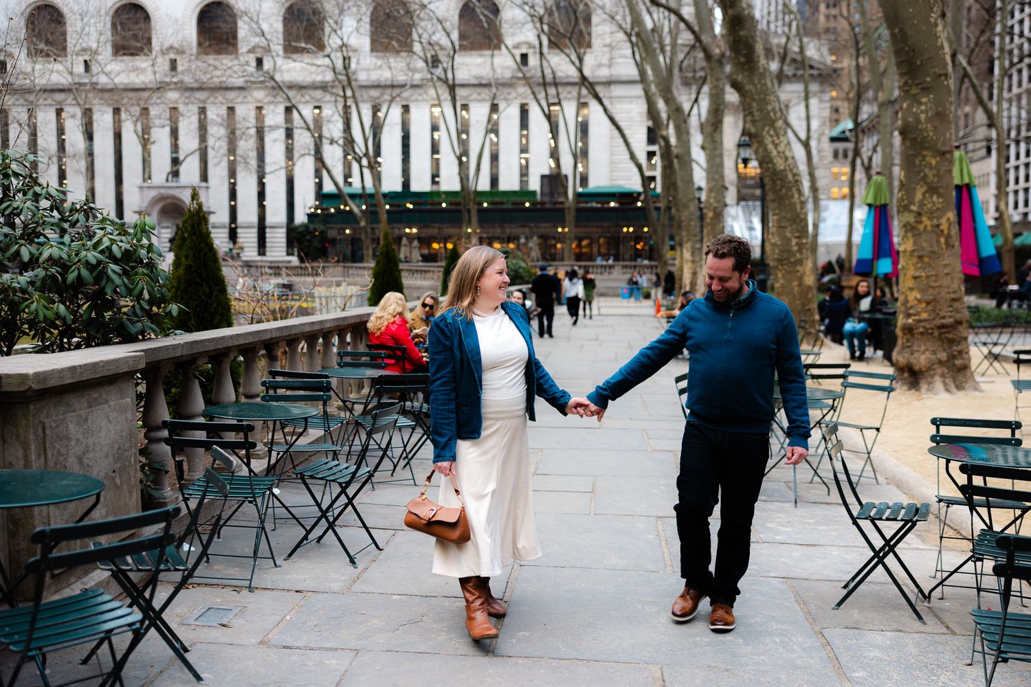 Couple holding hands walking through Bryant Park NYC with green chairs and historic building backdrop.