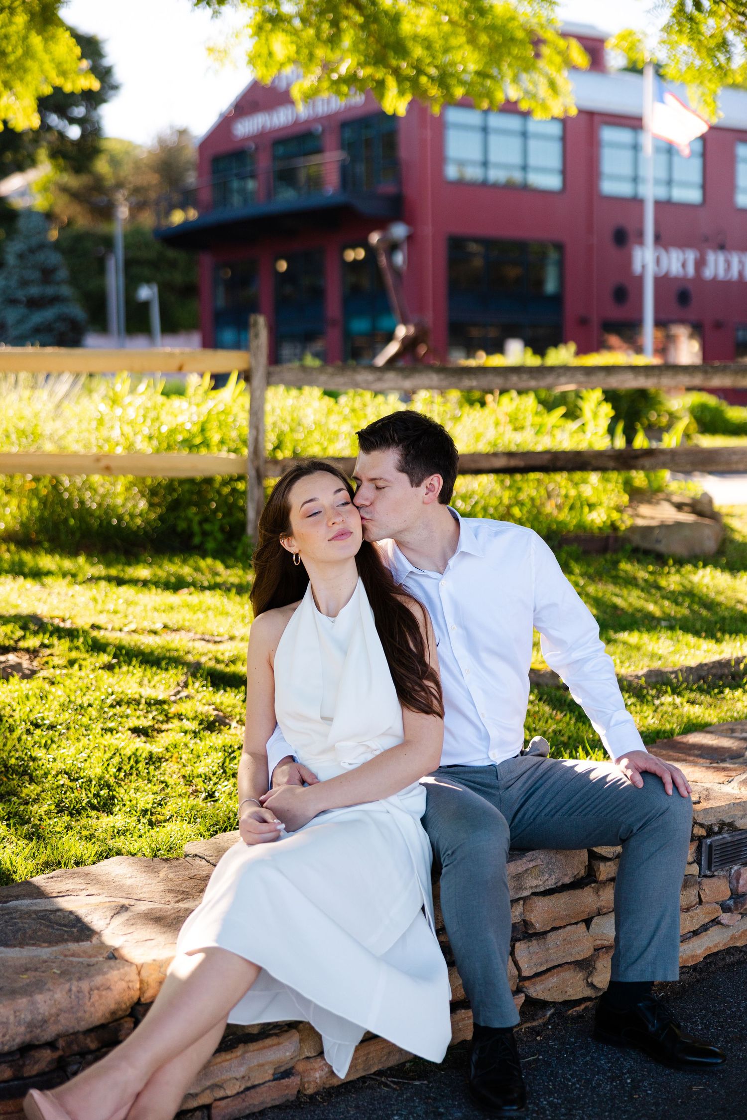 A couple sits on a stone wall outdoors, man in white shirt kissing woman in white dress on the cheek.