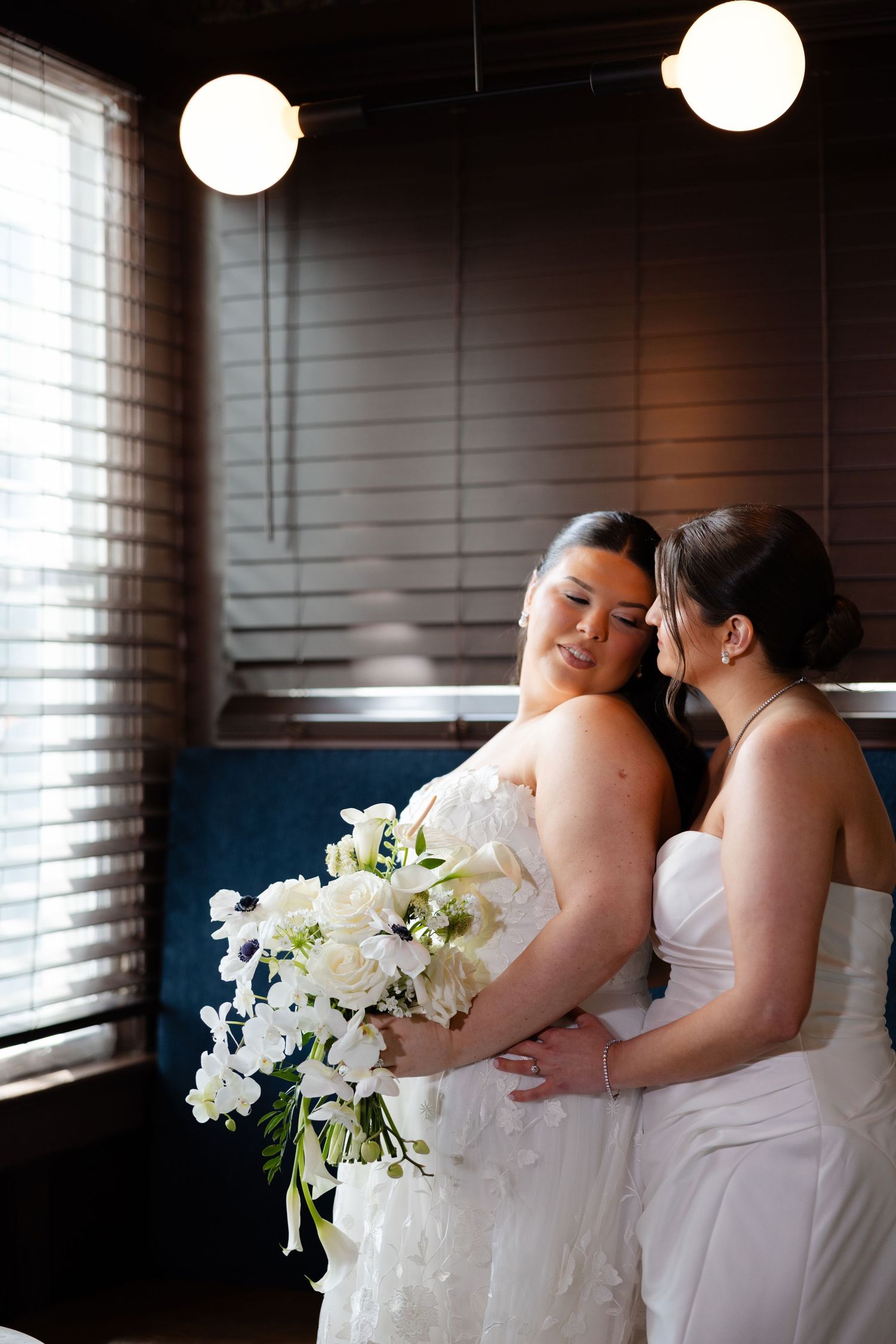 Two brides in white gowns share an intimate moment, one holding a white floral bouquet, in a warmly lit indoor setting.