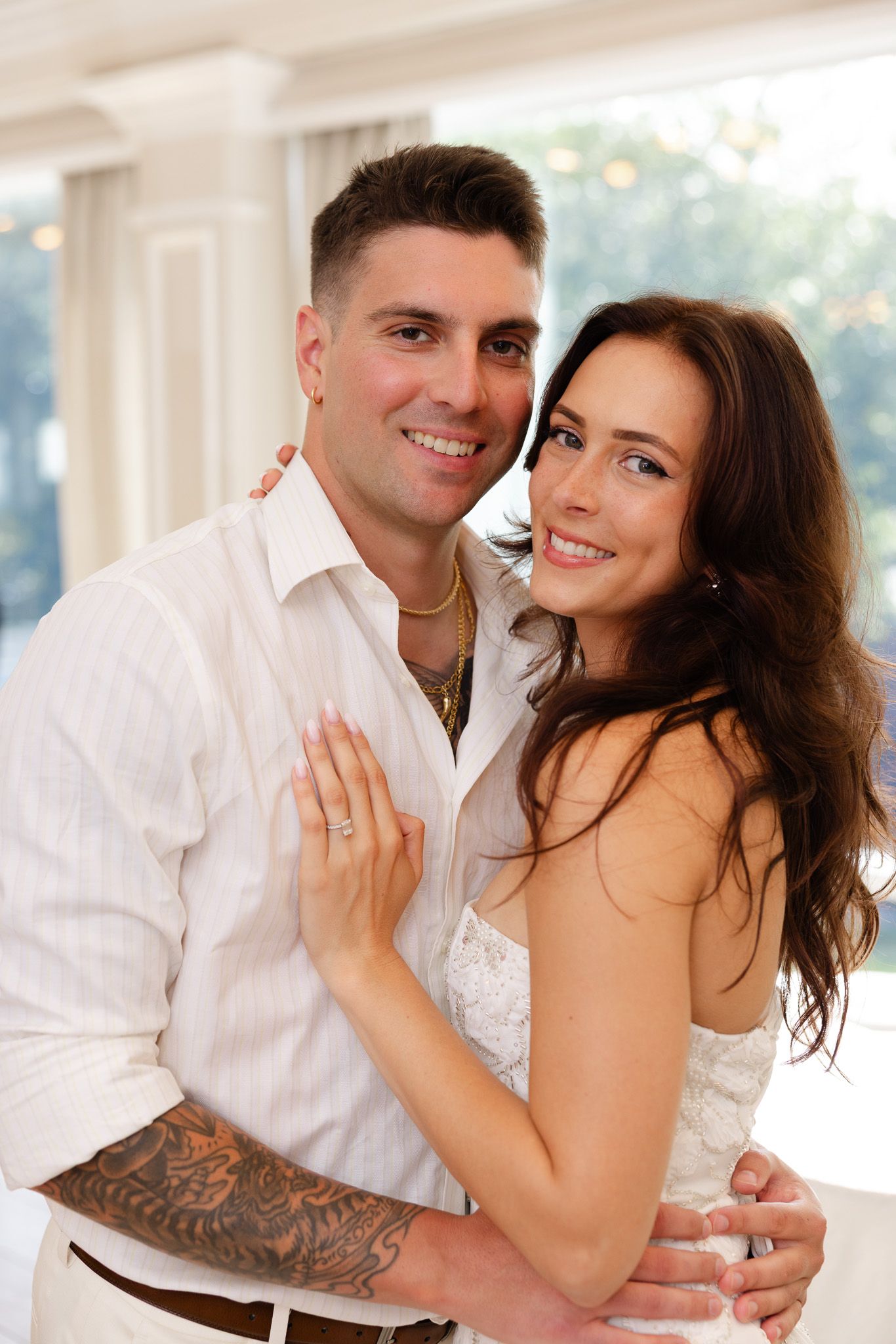 Happy newlywed couple embracing, bride showing wedding ring, groom with tattoos in white shirt.