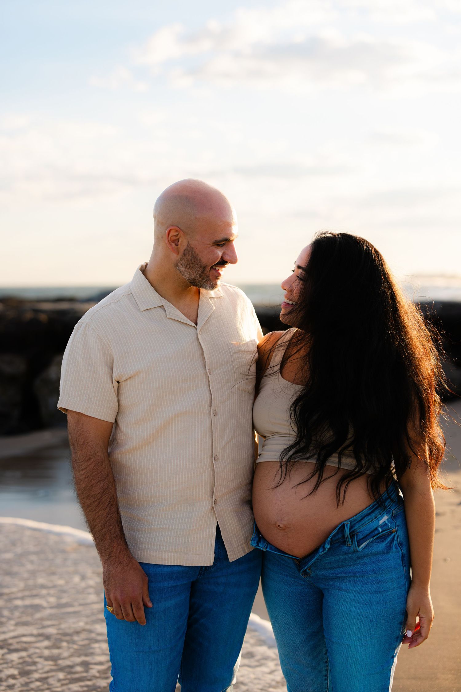 A bald man and pregnant woman smile at each other on a beach at sunset, both wearing jeans and light tops.