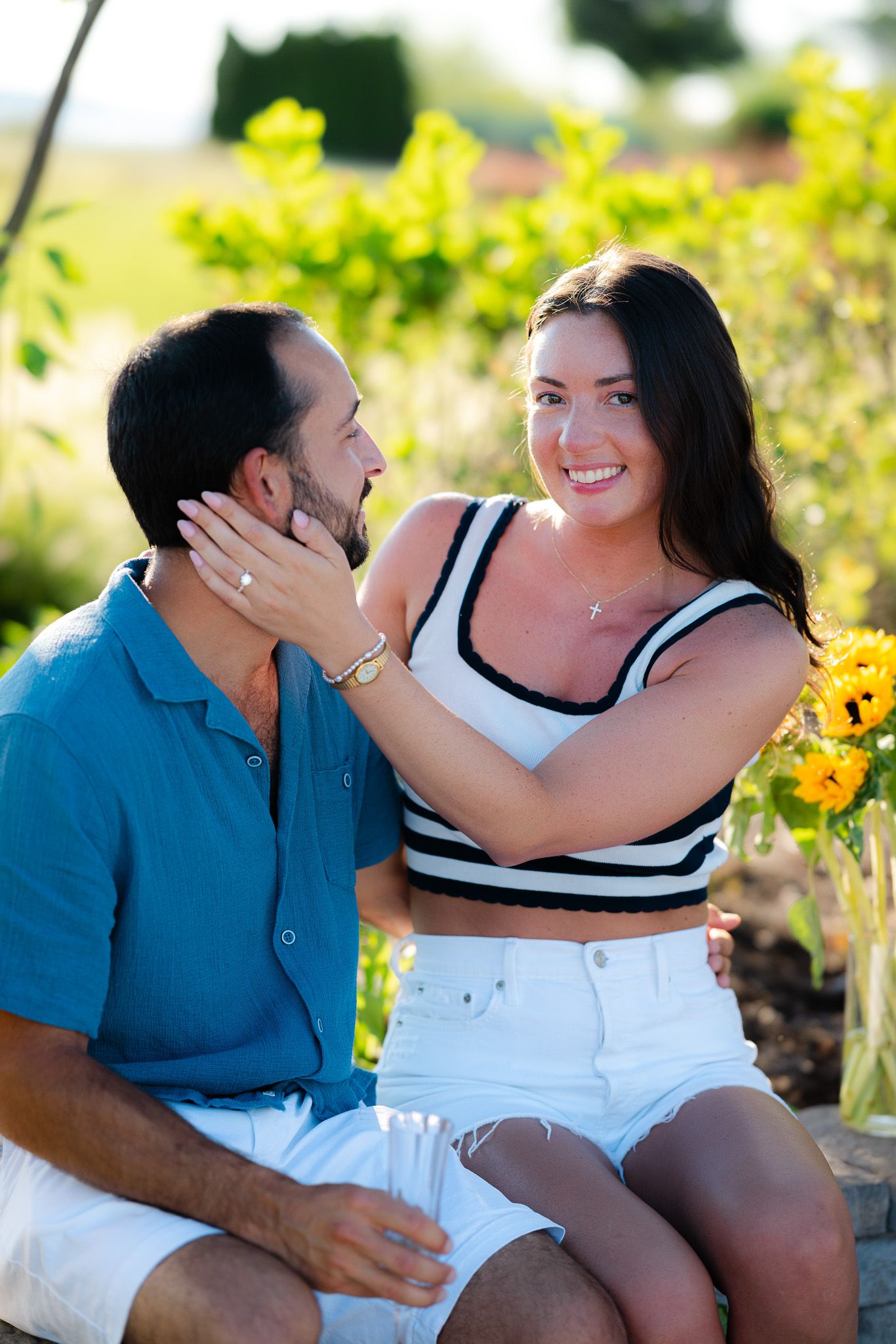 Smiling woman touching mans face while sitting together near sunflowers in a sunny garden.