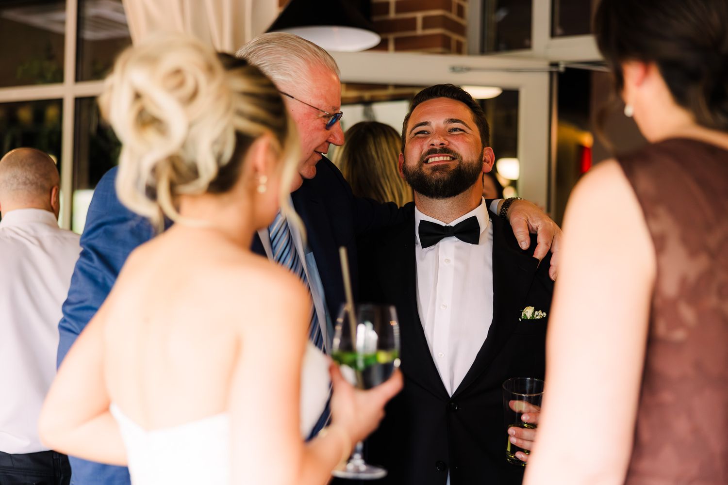 Groom in tuxedo with bow tie smiling and socializing with wedding guests during reception celebration.
