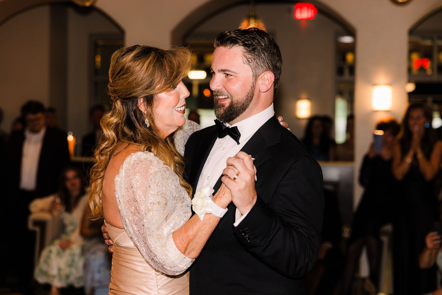 Couple shares first dance at wedding reception, groom in tuxedo and bride in lace dress, guests watching in background.