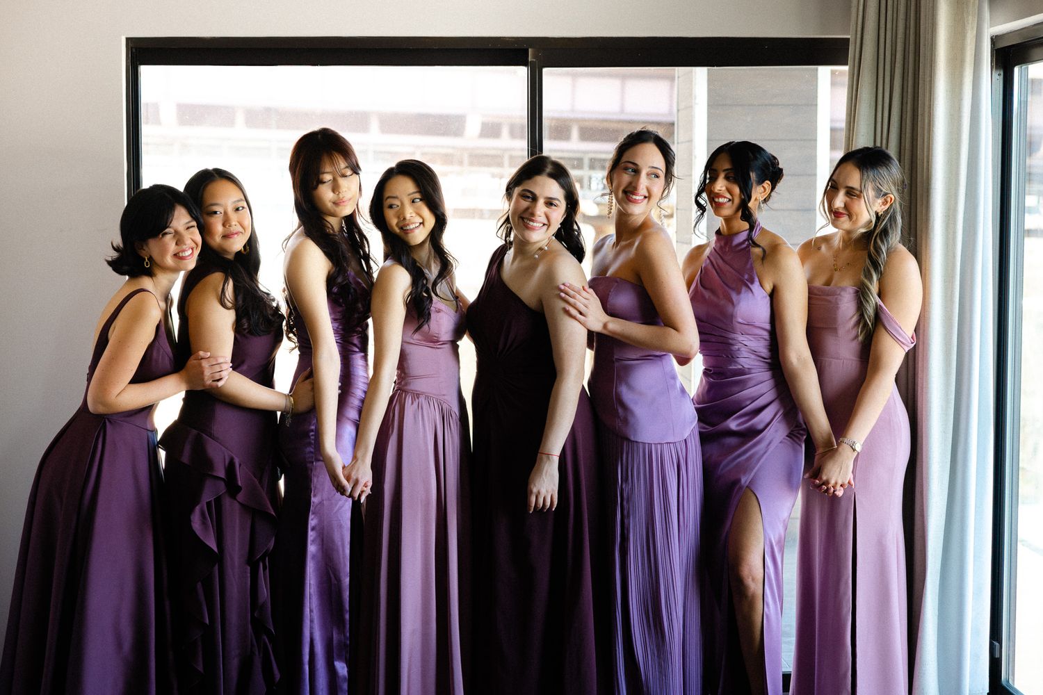 Eight bridesmaids in purple gowns pose together smiling in a bright hotel room before a wedding ceremony.
