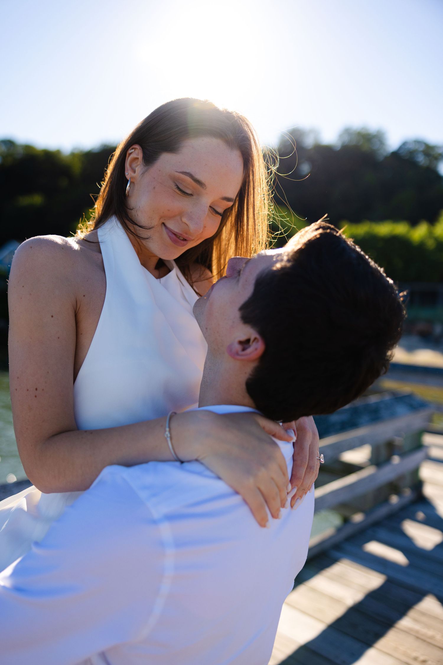 Couple in white embracing on a sunny dock, sharing a romantic moment with golden backlight and lush greenery.