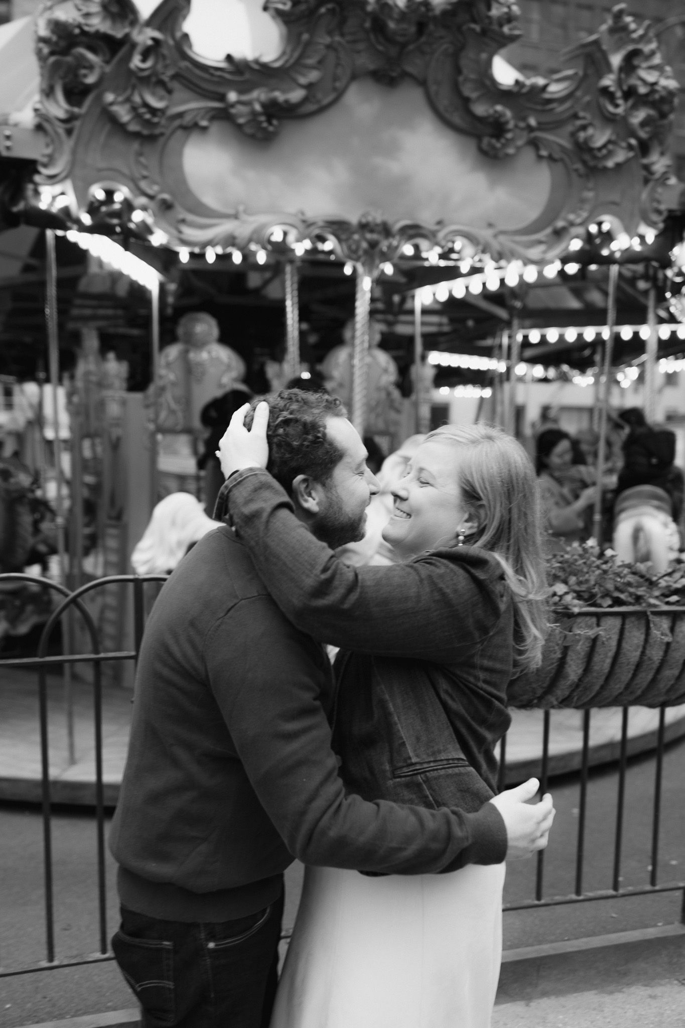 Black and white photo of a couple embracing and kissing in front of a lit carousel at an amusement park.