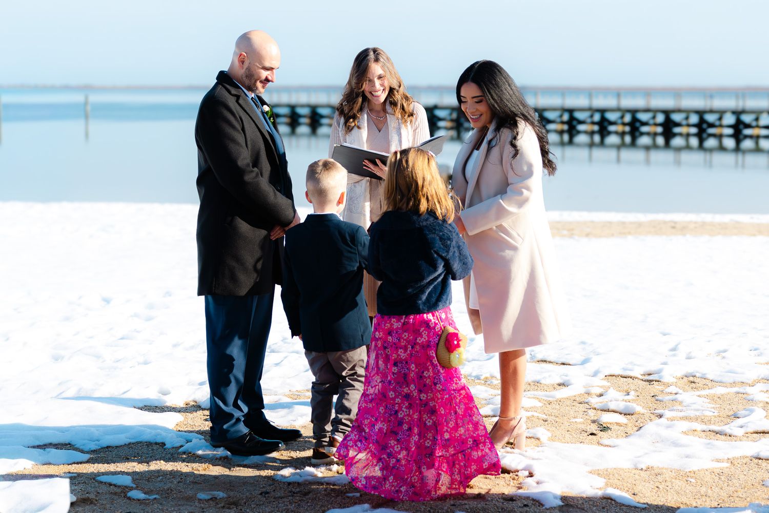 Winter beach wedding ceremony with couple, officiant, and two children in snow near a pier.