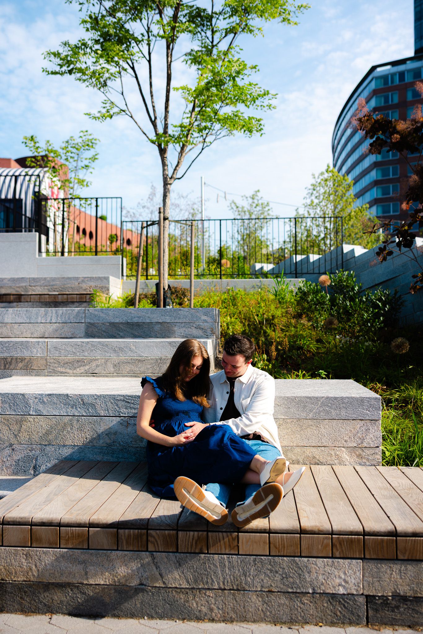 Couple sitting closely on stone steps in a modern park, surrounded by greenery and contemporary buildings.