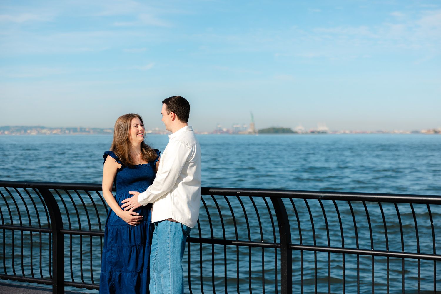 Couple embracing by waterfront railing with Statue of Liberty in background on sunny day.