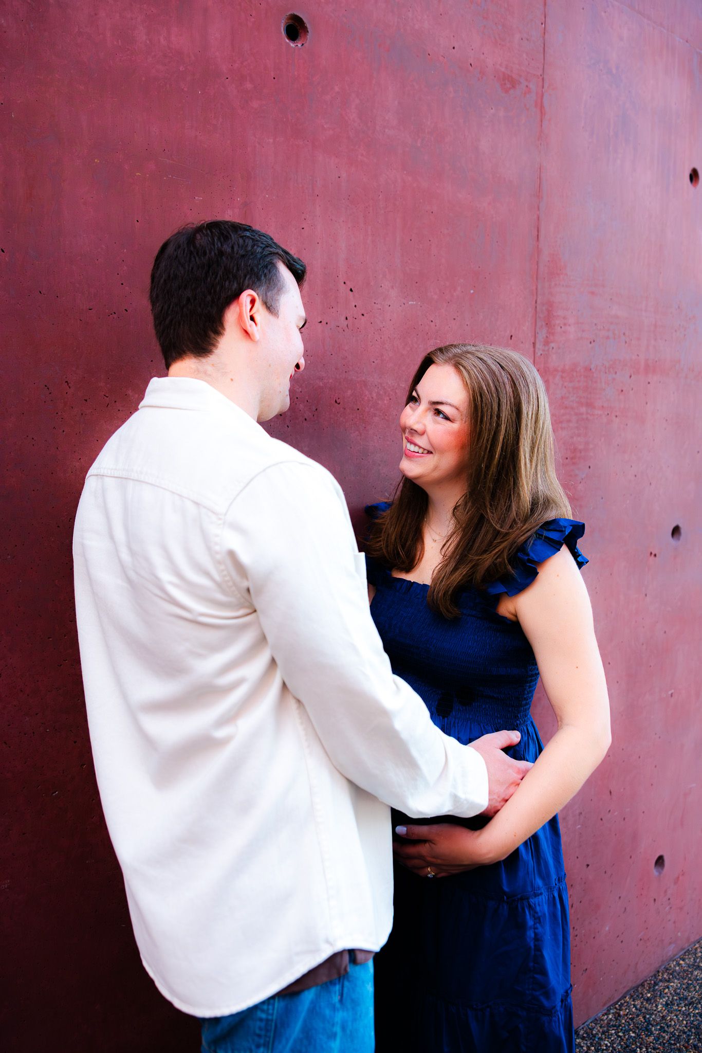 A smiling couple in a romantic embrace against a red wall, woman in blue dress and man in white shirt.