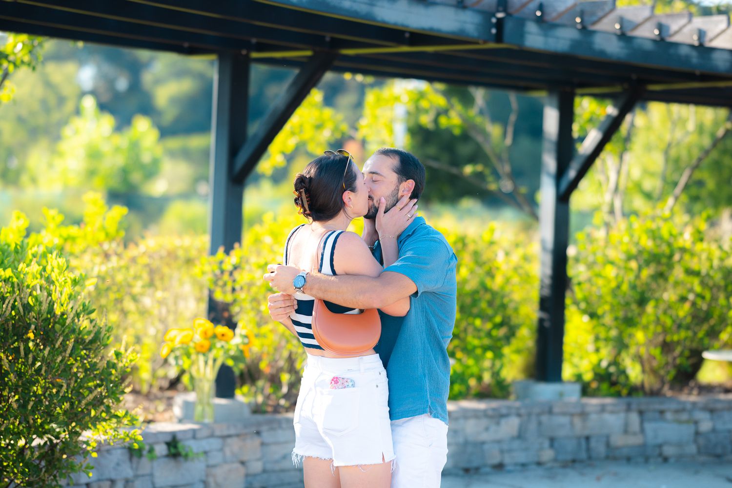 A couple shares a romantic kiss outdoors under a pergola surrounded by lush green trees on a sunny day.