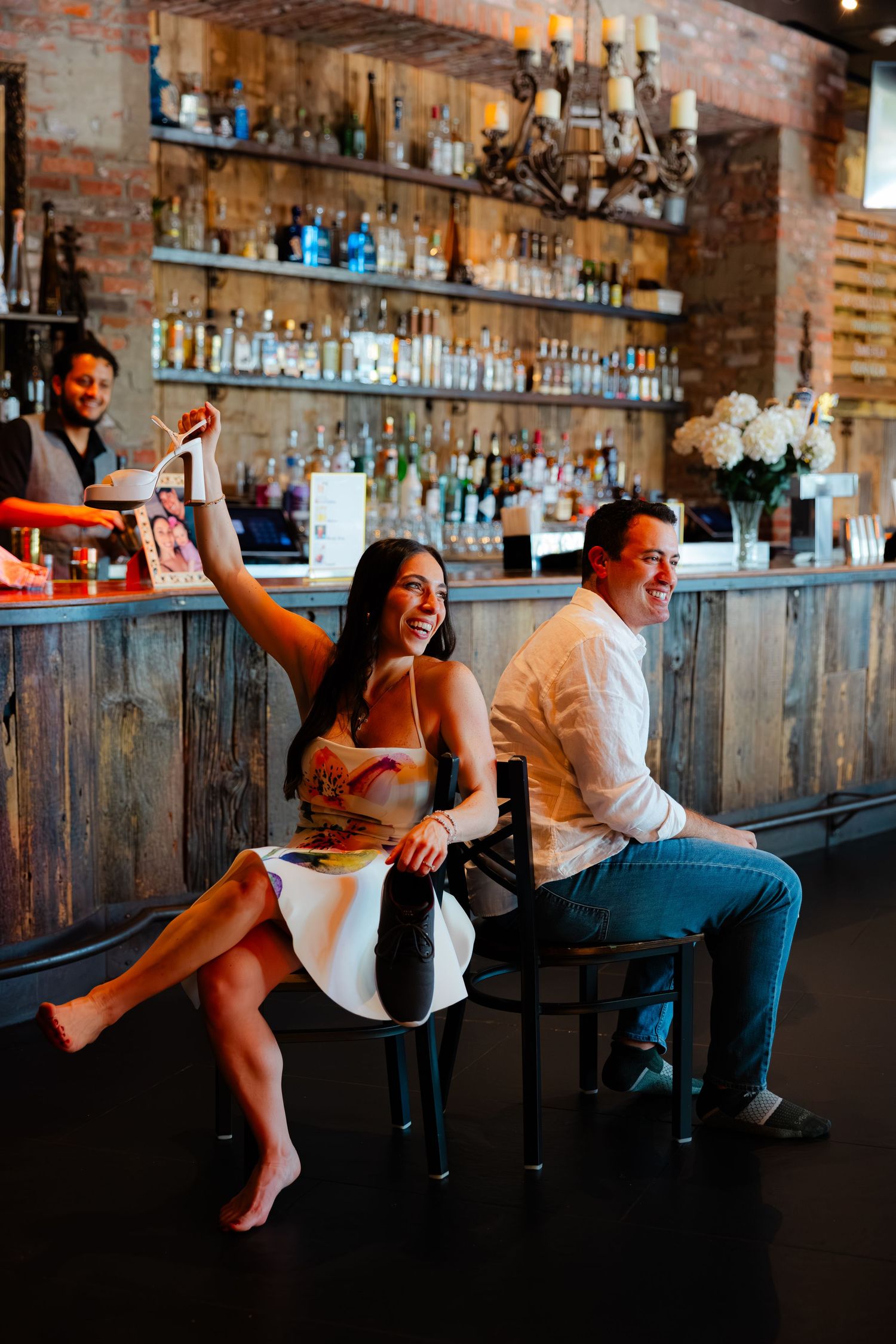A joyful couple celebrates at a rustic bar, woman raising a drink and laughing, man smiling beside her.