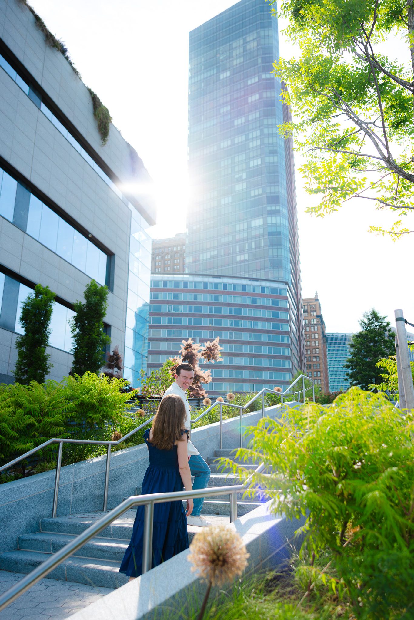 Couple in blue attire on urban garden steps with sunlit glass skyscrapers and lush greenery in the background.