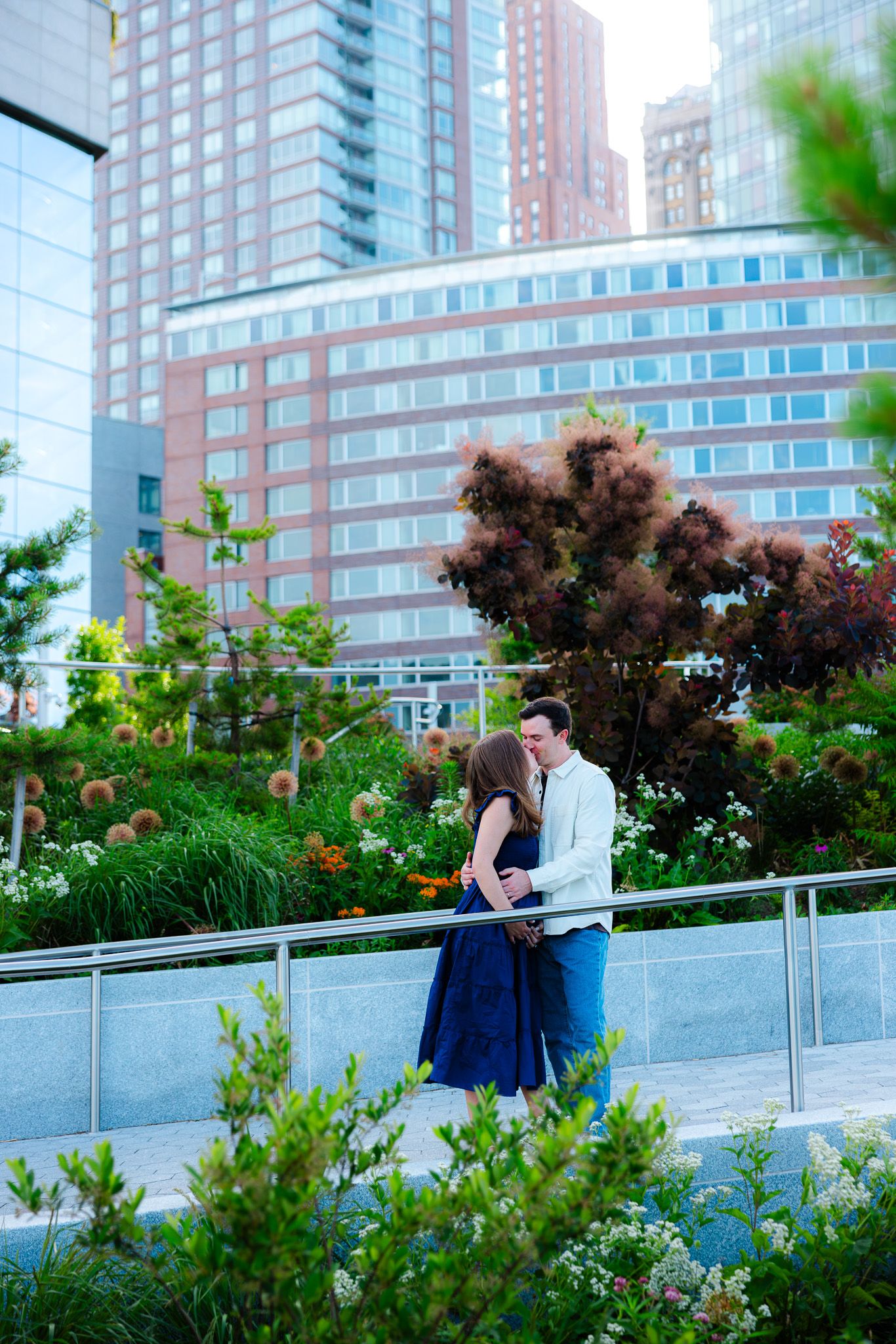 Couple sharing a romantic moment in an urban garden with lush greenery and modern city buildings in the background.