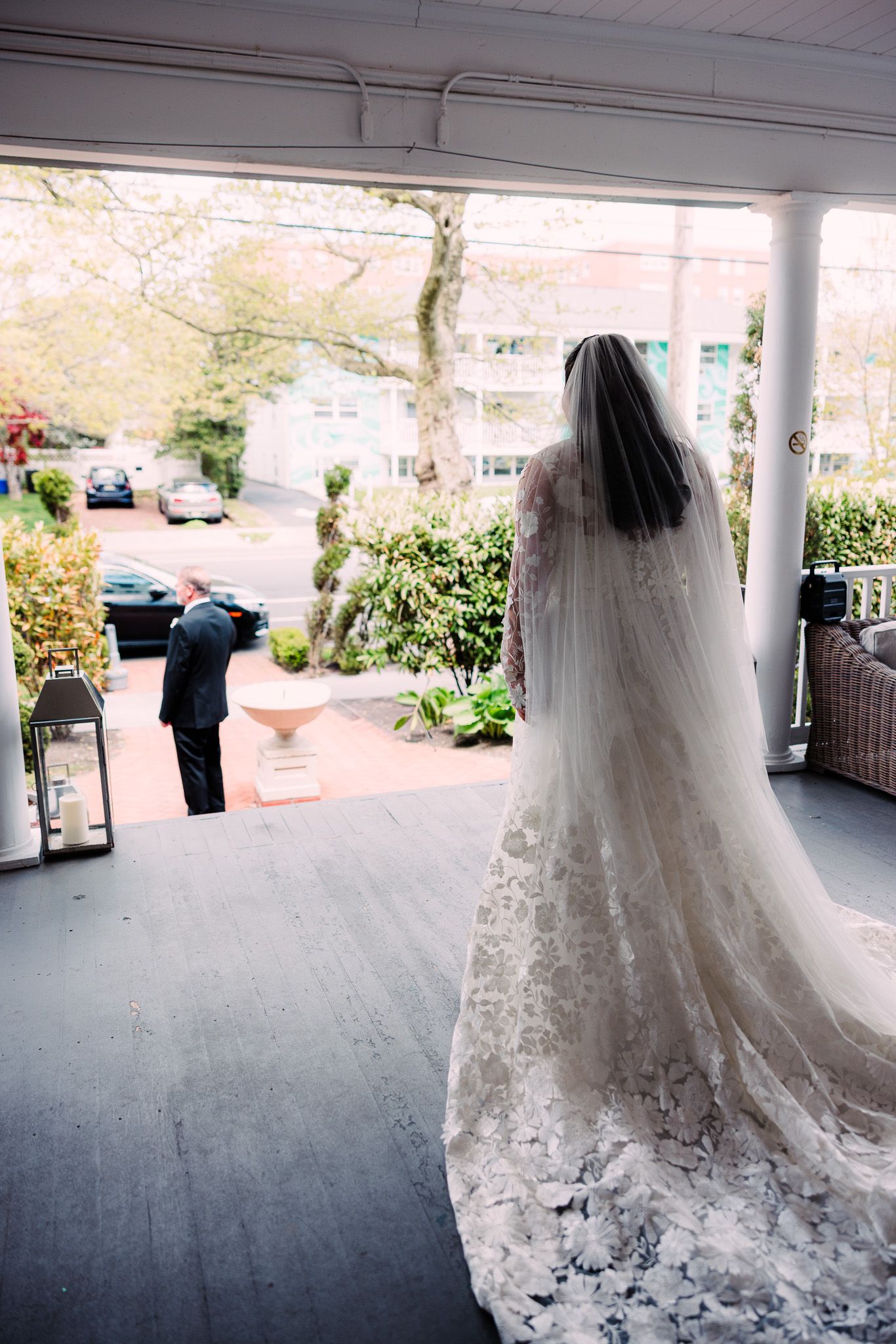 Bride in lace gown and veil stands on porch as groom turns away for first look at outdoor wedding venue.