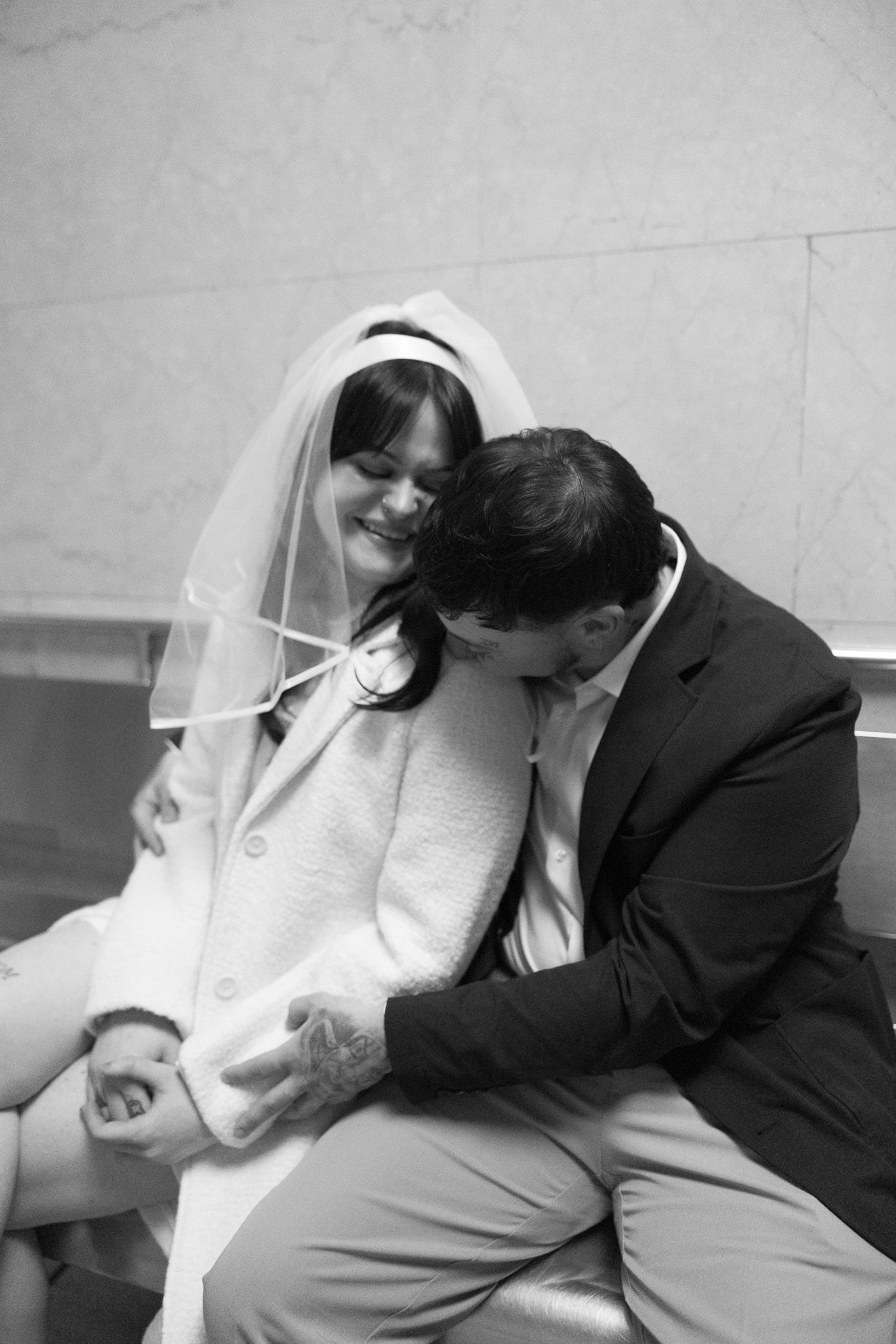 Black and white photo of a smiling bride in veil and white coat with groom in suit sitting together on wedding day.