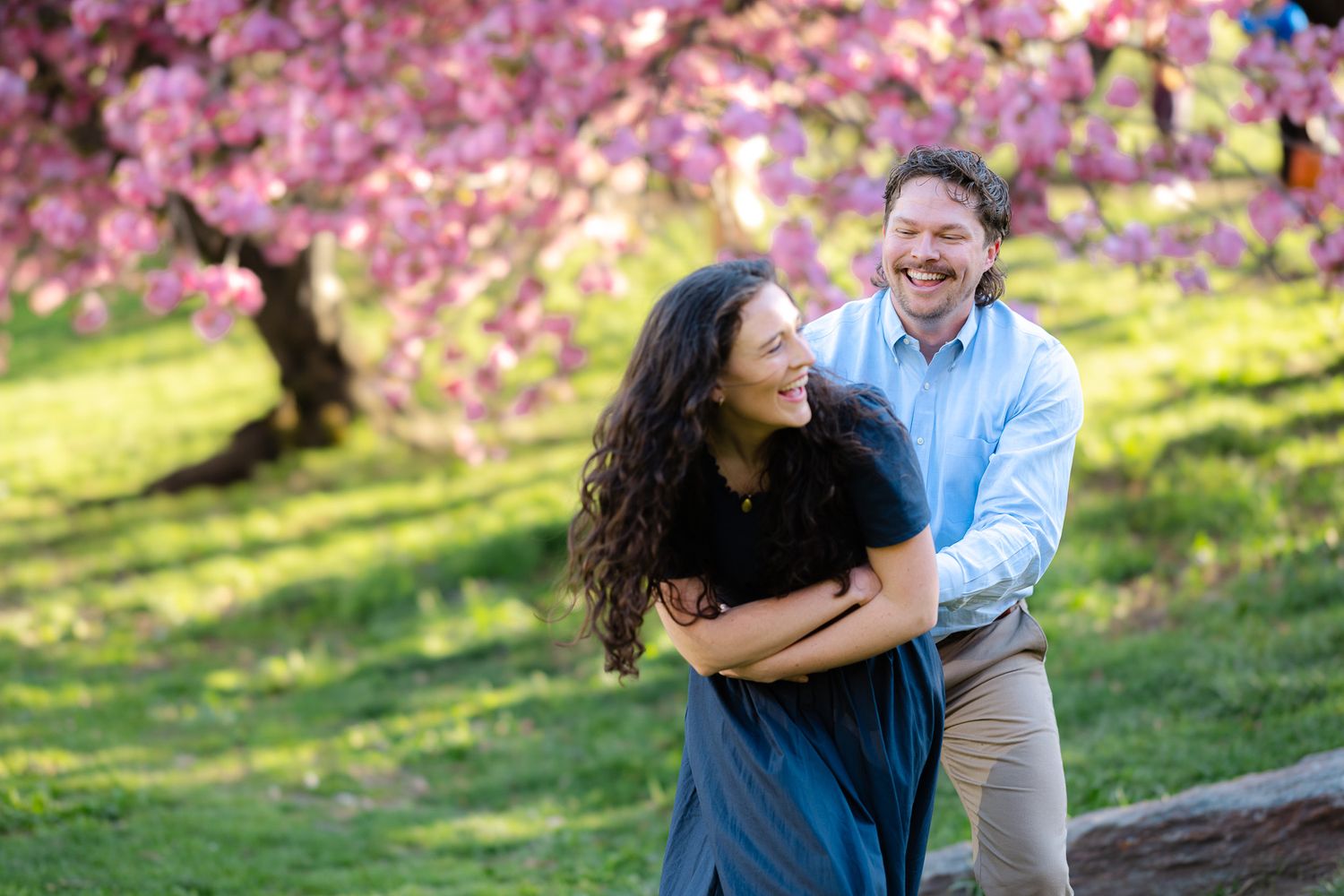 Laughing couple embracing outdoors with vibrant pink cherry blossom trees in the background.