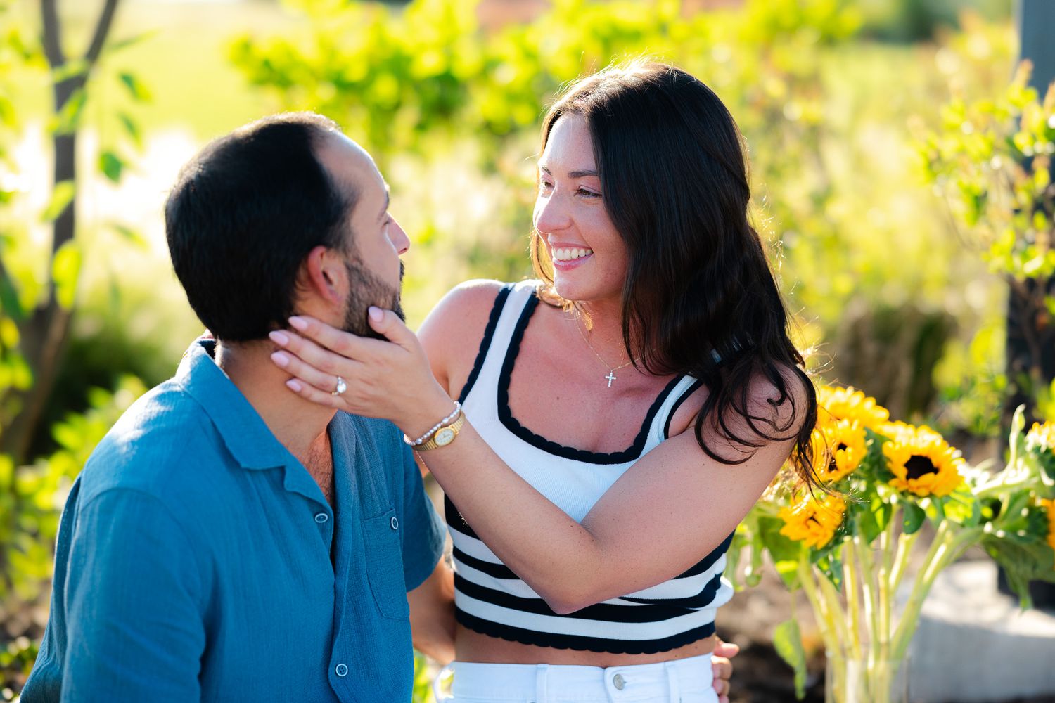 Happy couple gazing at each other lovingly outdoors with sunflowers in background, woman smiling brightly.