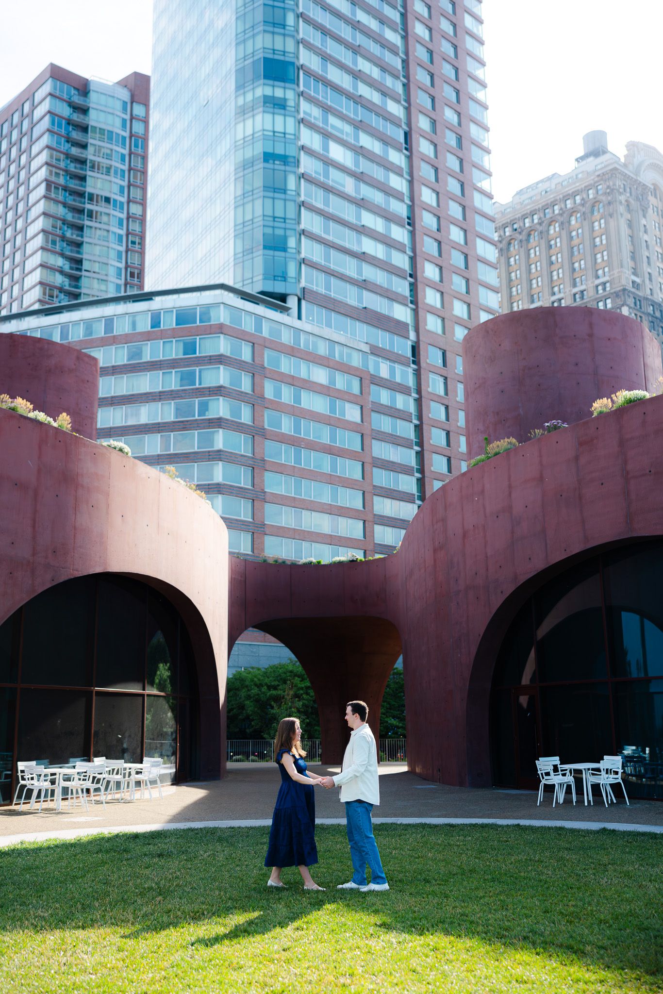 Couple holding hands on green lawn before red arched sculpture with city skyscrapers in background.