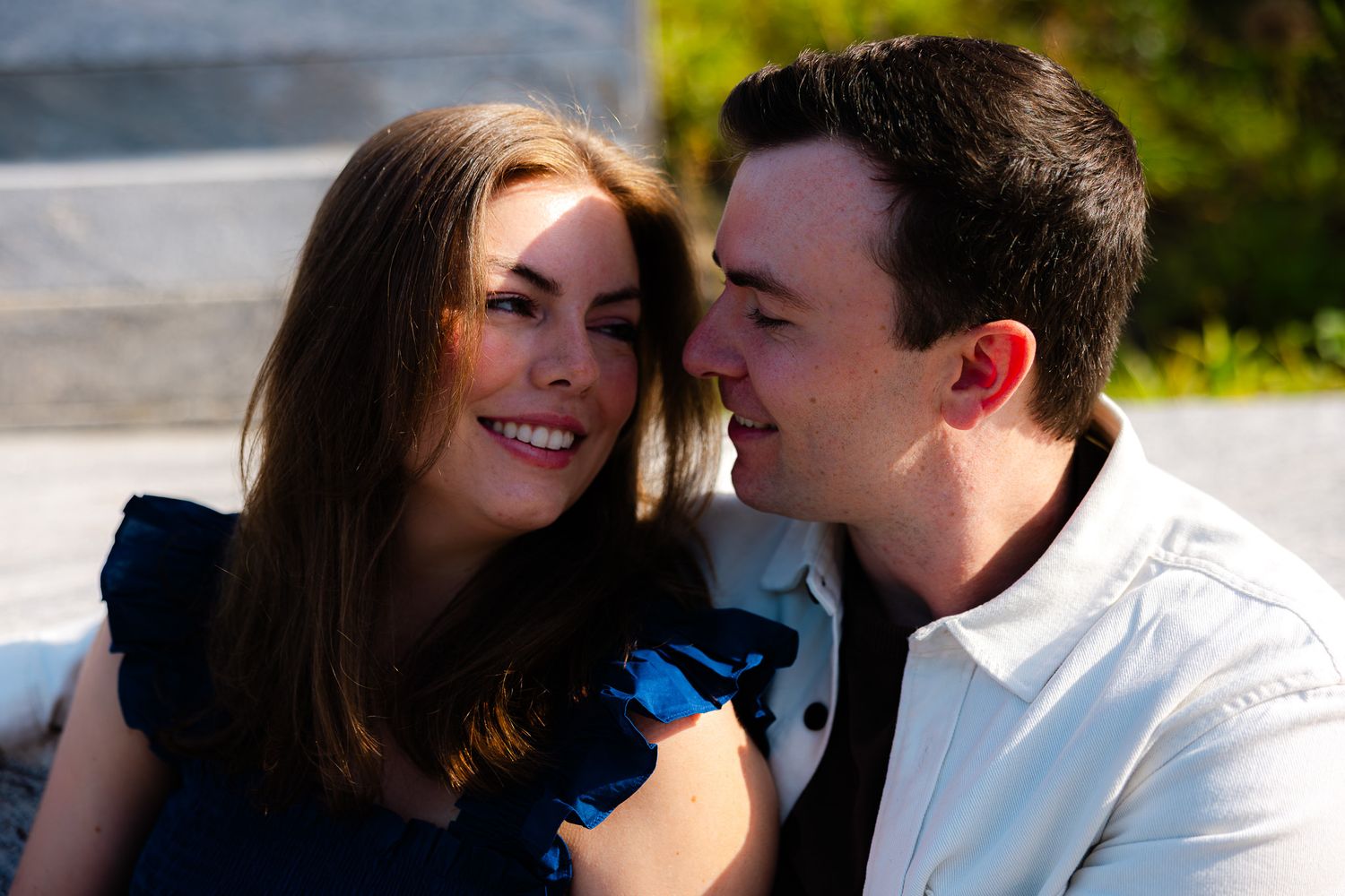 A smiling couple sitting close together outdoors, the woman in a blue top and the man in a white shirt, sharing a joyful moment.