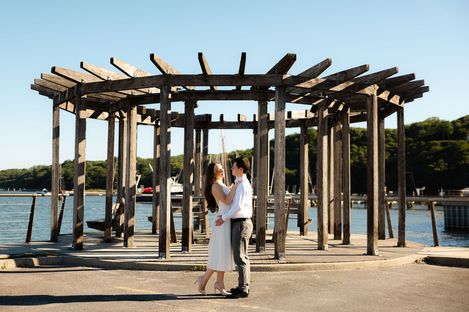Couple embracing under a wooden pergola on a waterfront dock on a sunny day.