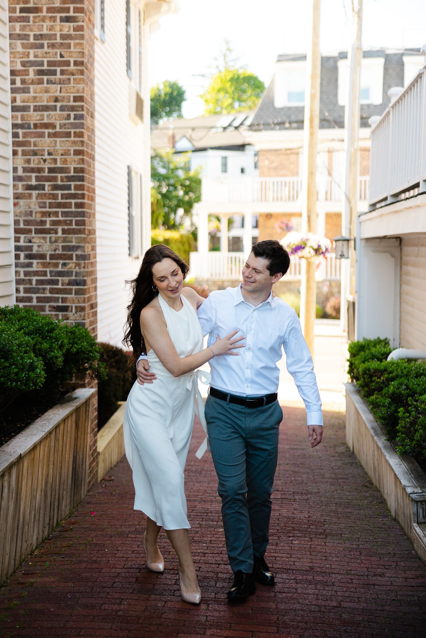 A couple walks arm in arm down a brick pathway between buildings, woman in white dress, man in blue shirt and teal pants.