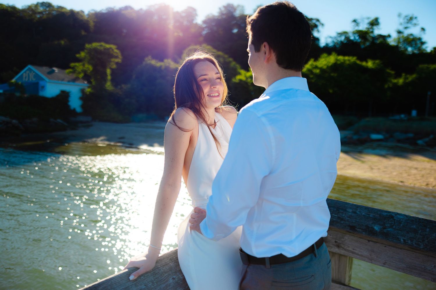 Couple sharing a romantic moment on a sunny pier by the beach, woman in white dress smiling warmly at her partner.