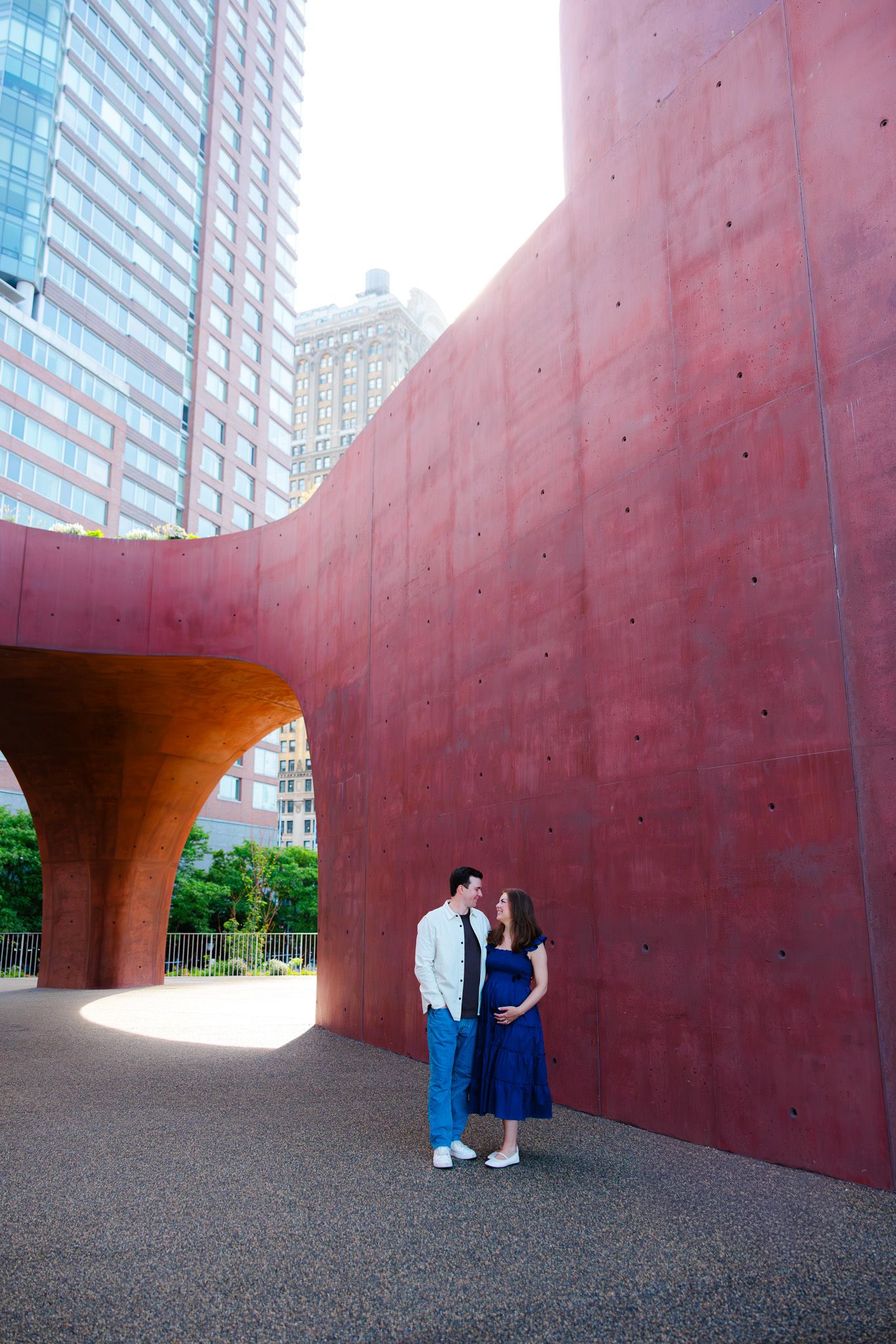 Couple embracing near a large red concrete sculpture in an urban park with city skyscrapers in the background.