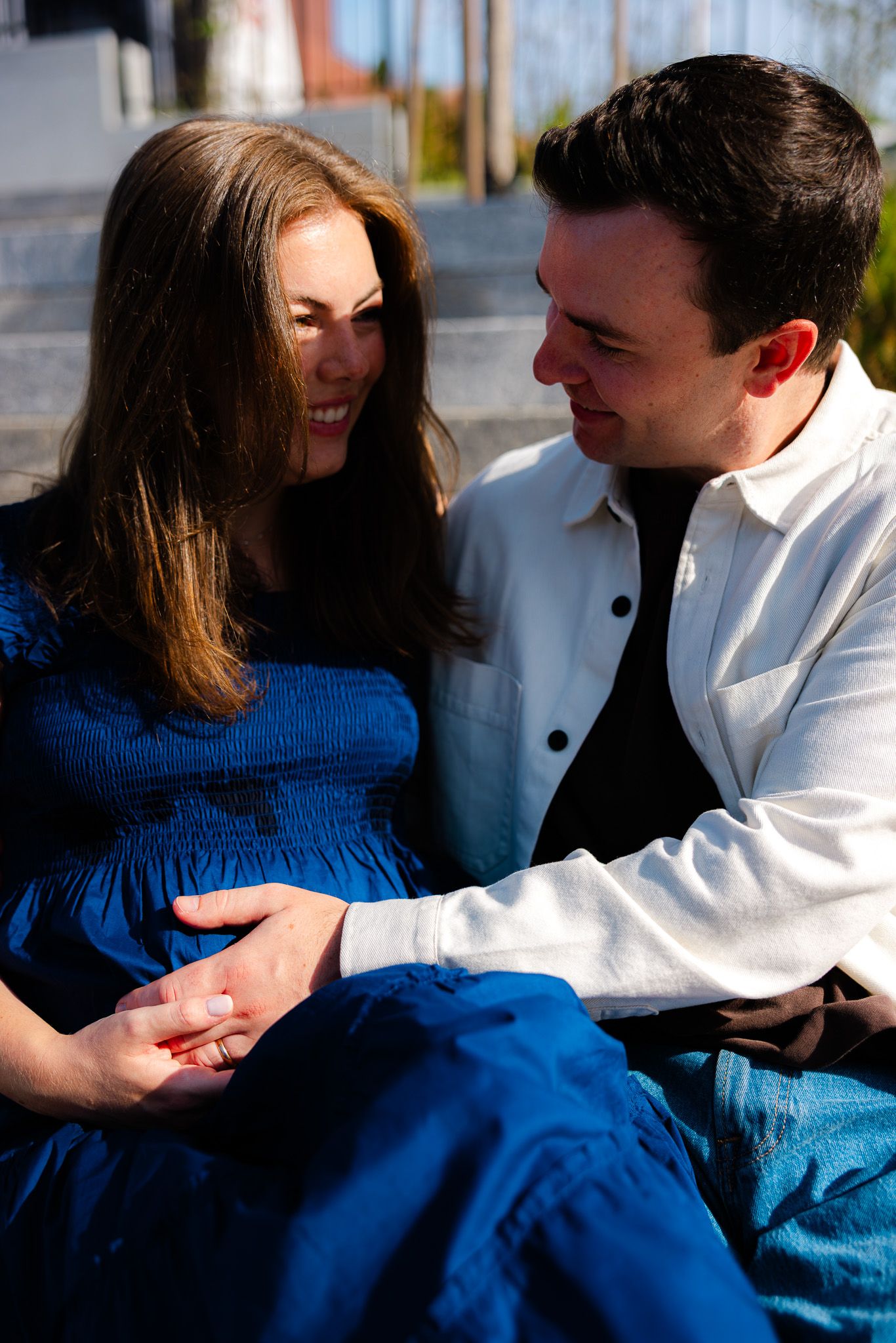 Happy couple sitting outdoors, man in white jacket holding pregnant womans hand on her baby bump.