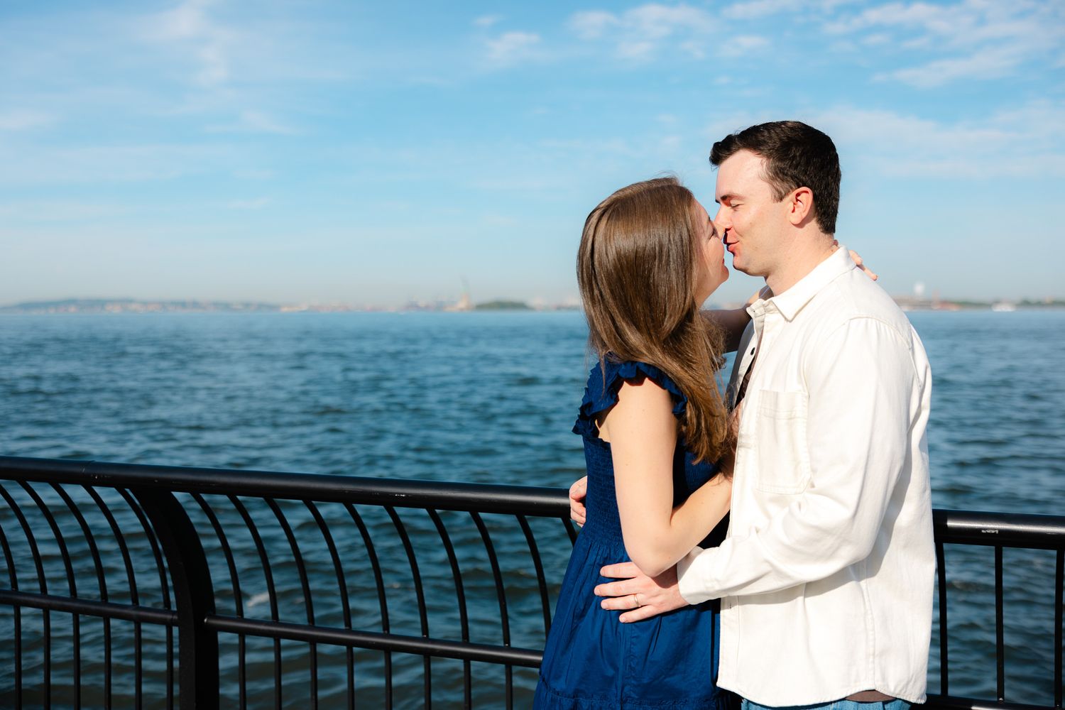 Couple sharing a romantic kiss by a waterfront railing with blue sky and ocean in the background.