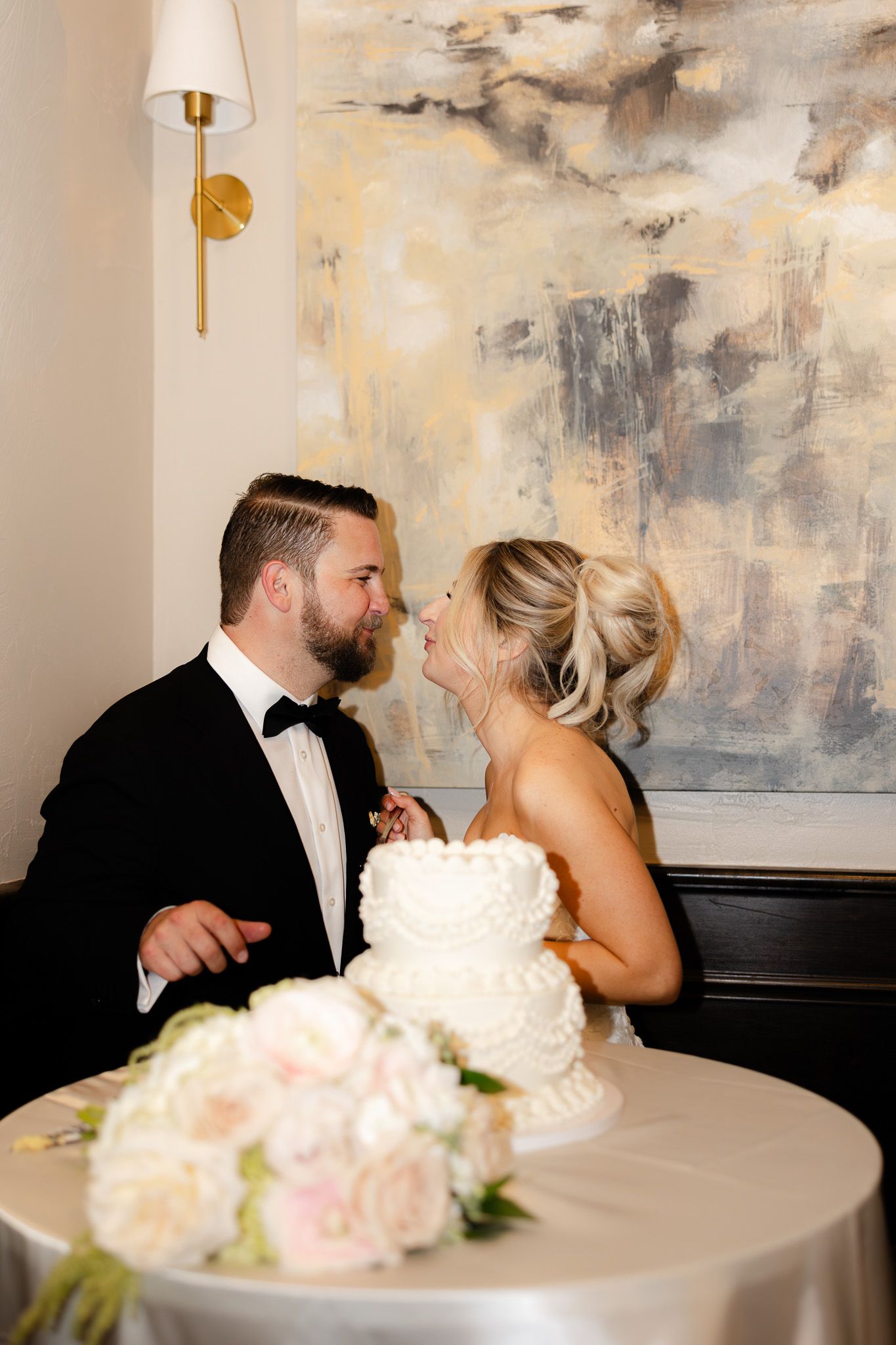Bride and groom share a romantic moment by their white wedding cake beneath an abstract painting.