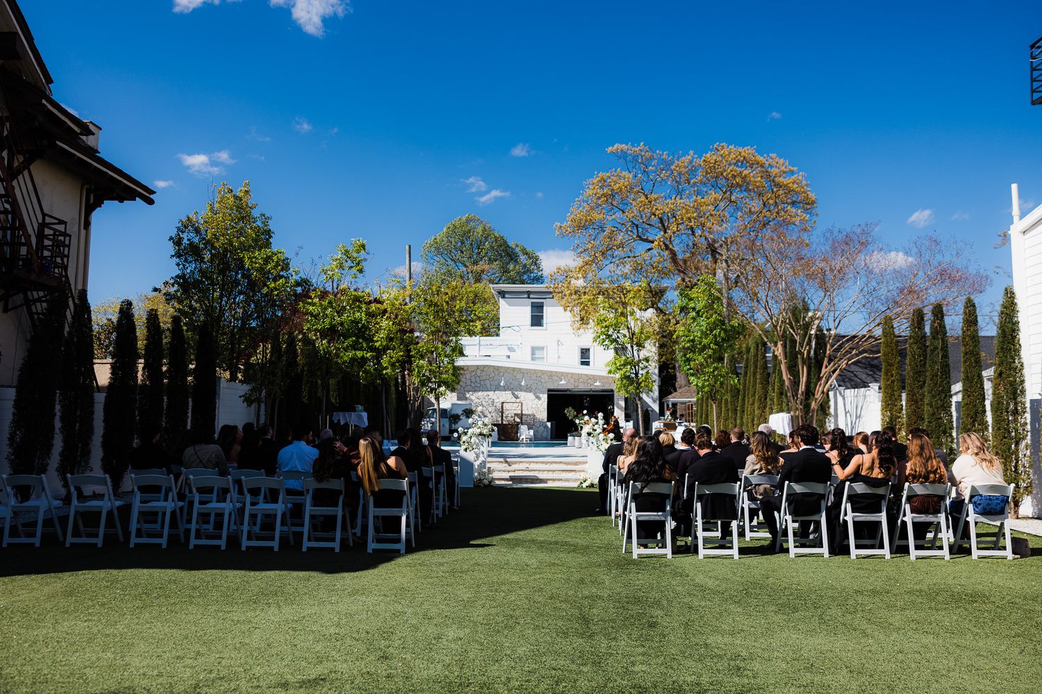Guests seated in white chairs on a sunny lawn for an elegant outdoor wedding ceremony.