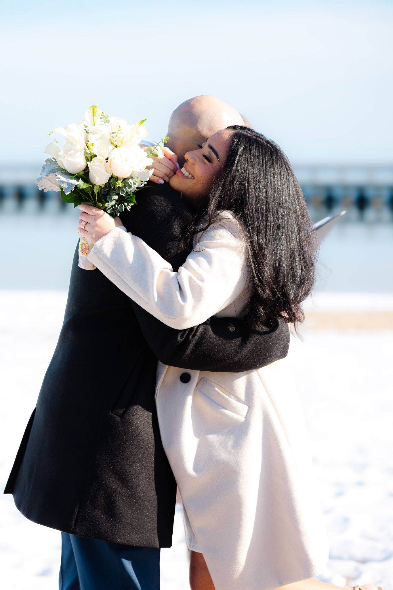 Couple embracing on a snowy beach, woman holding white roses bouquet, wearing elegant coats.