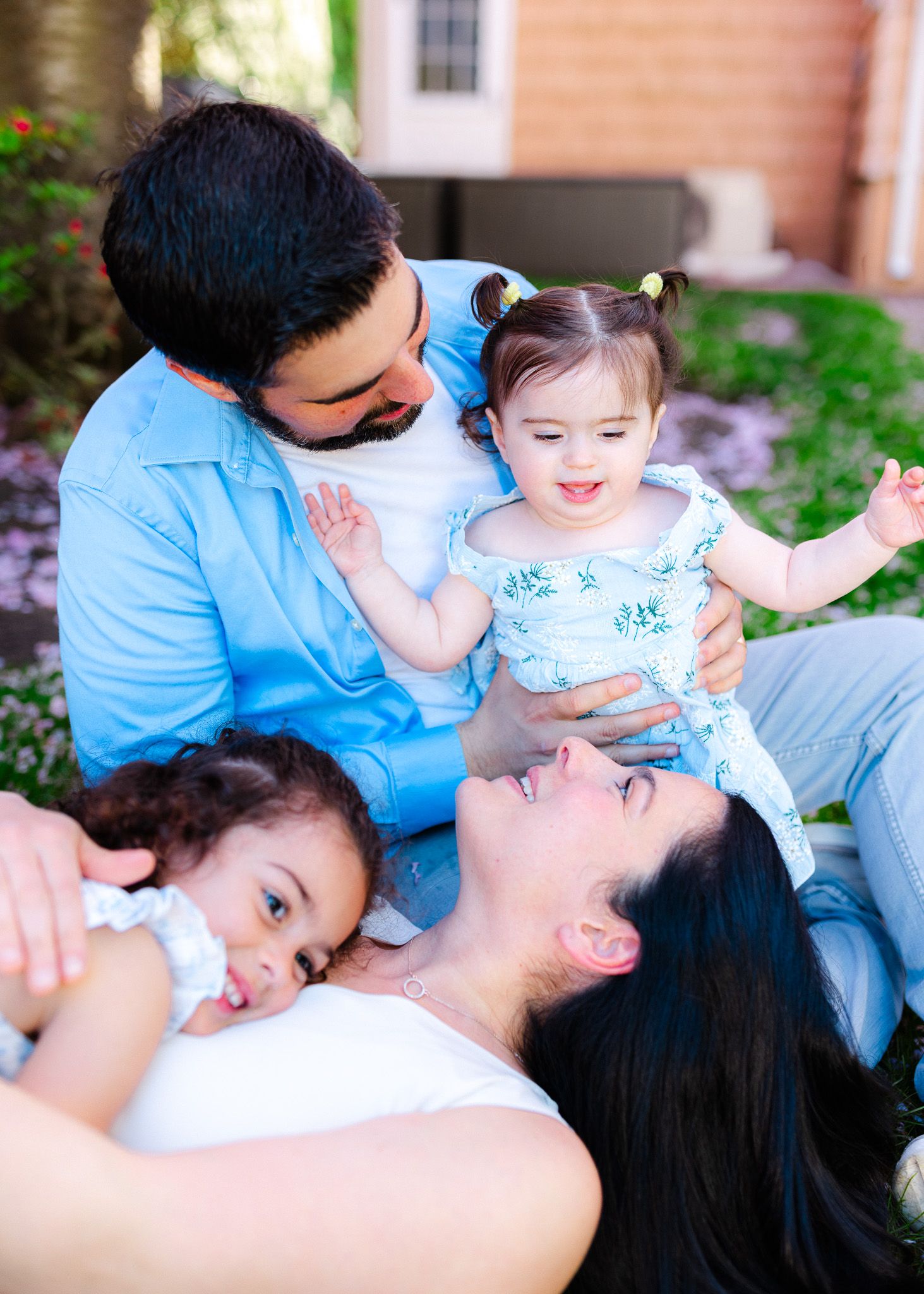 Happy family with father in blue shirt and mother lying on grass, playing with two young daughters outdoors.