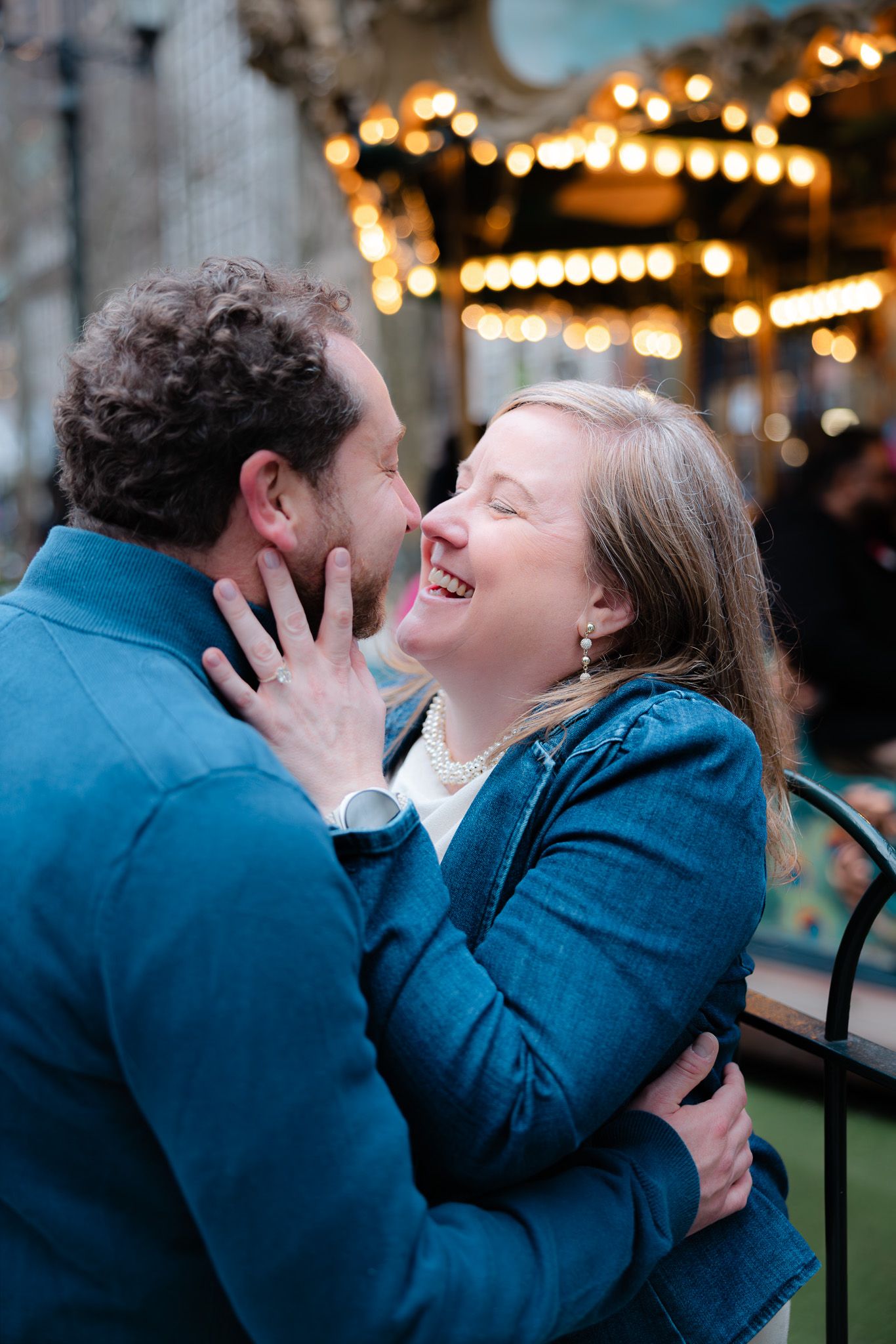 A joyful couple embracing and laughing together near a carousel with warm glowing lights in the background.