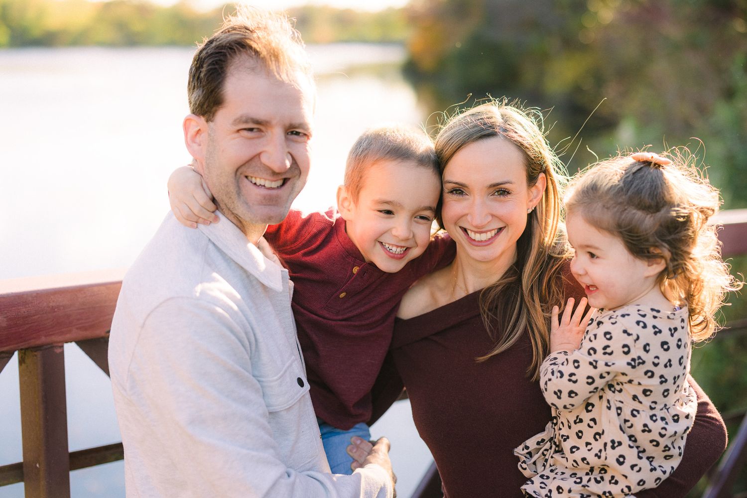 Happy family of four smiling together outdoors near a lake, parents holding young children in warm autumn light.