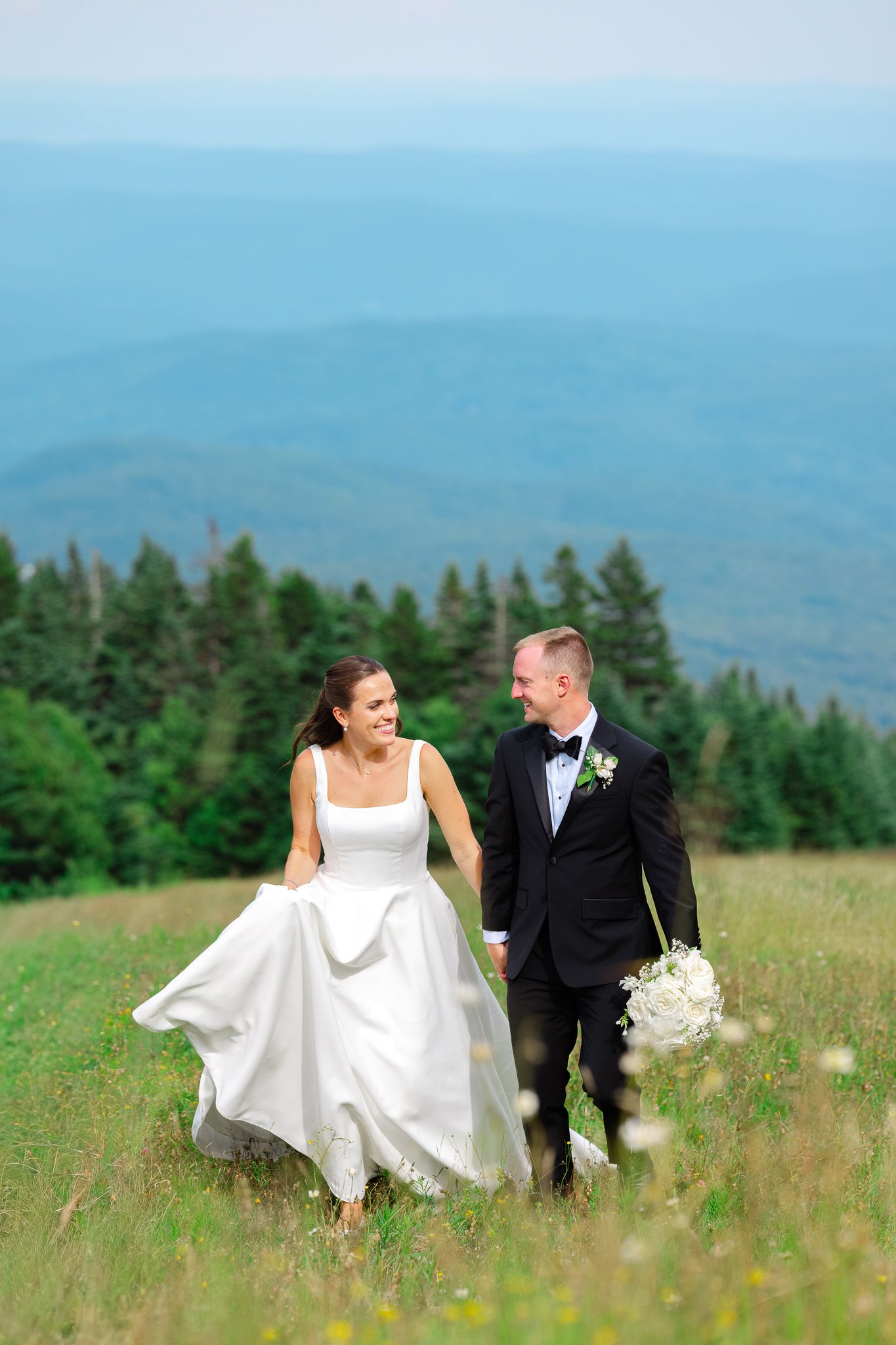 A bride and groom walk hand-in-hand through a mountain meadow, smiling at each other on their wedding day.