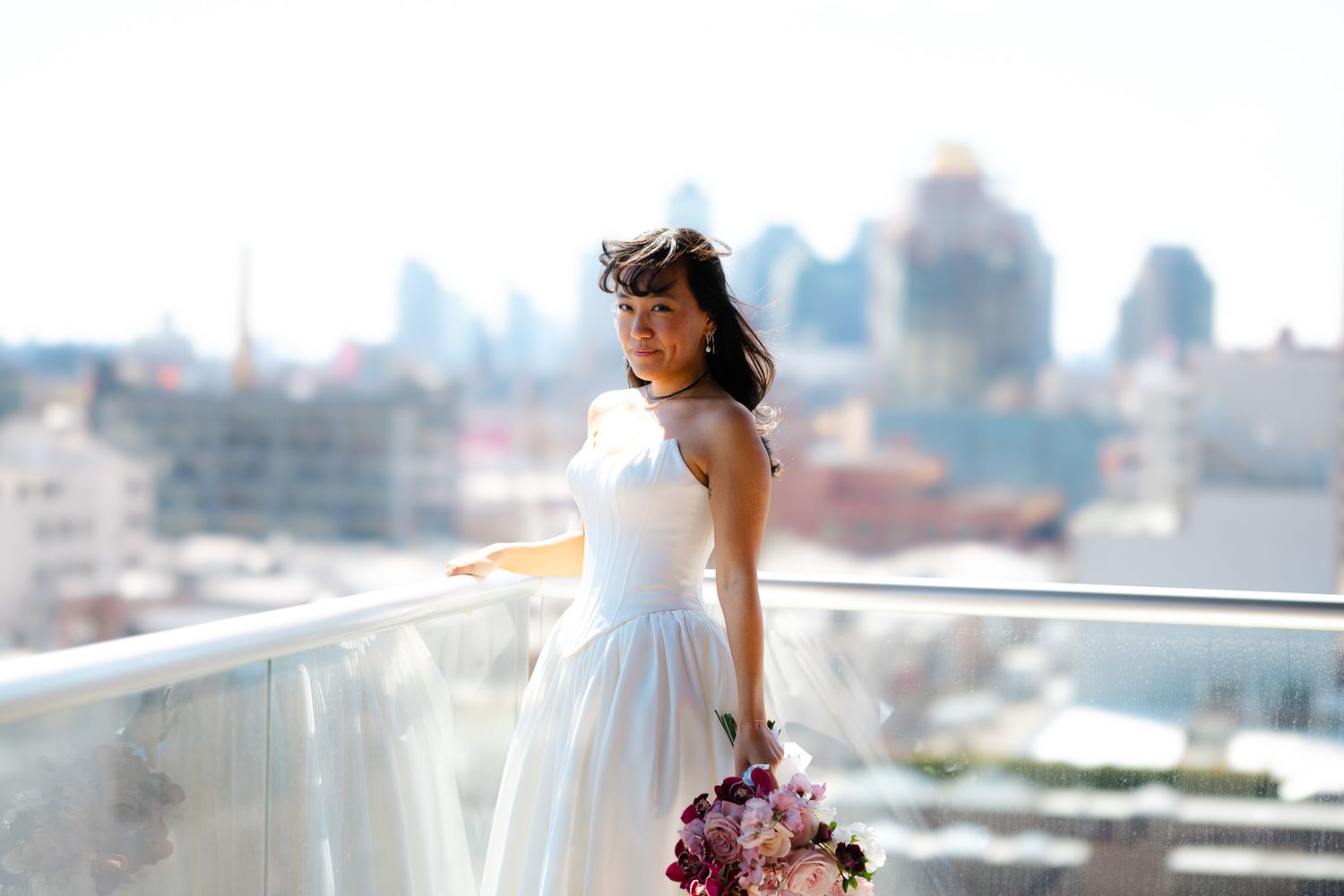 Bride in white dress holding a bouquet on a rooftop with a city skyline behind her.