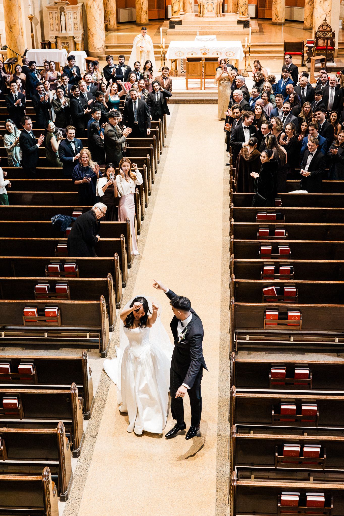 Bride and groom celebrate walking down the aisle of an ornate church surrounded by cheering wedding guests.