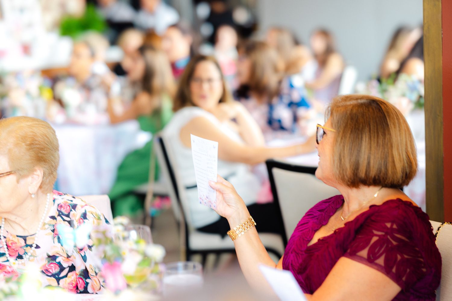 Women socializing at an elegant indoor event, with guests seated at flower-decorated tables in a bright venue.