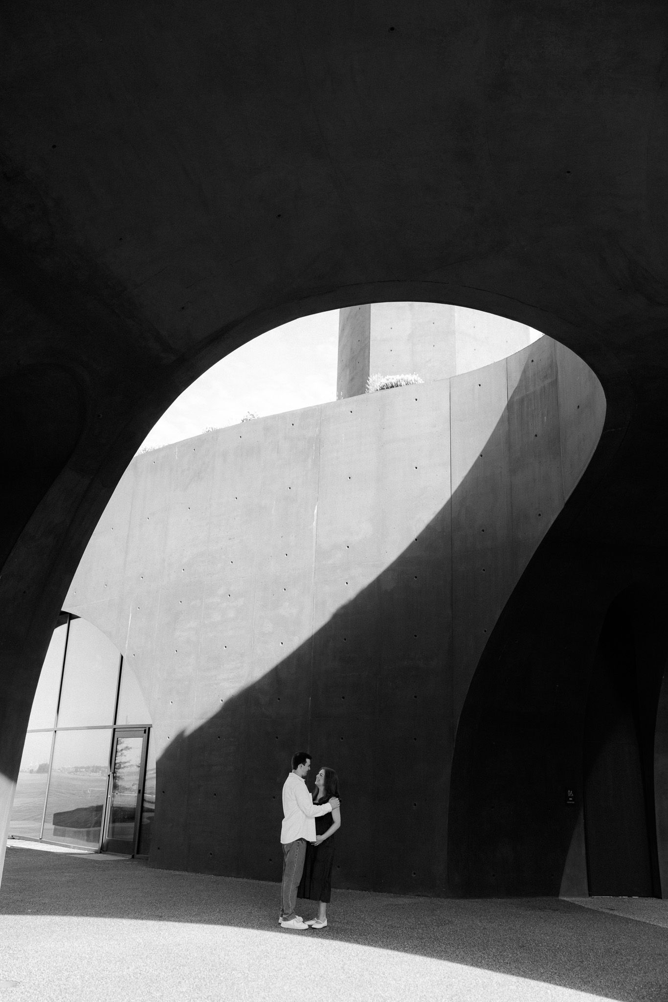 A couple embraces under a dramatic concrete arch in a striking black and white architectural photo.