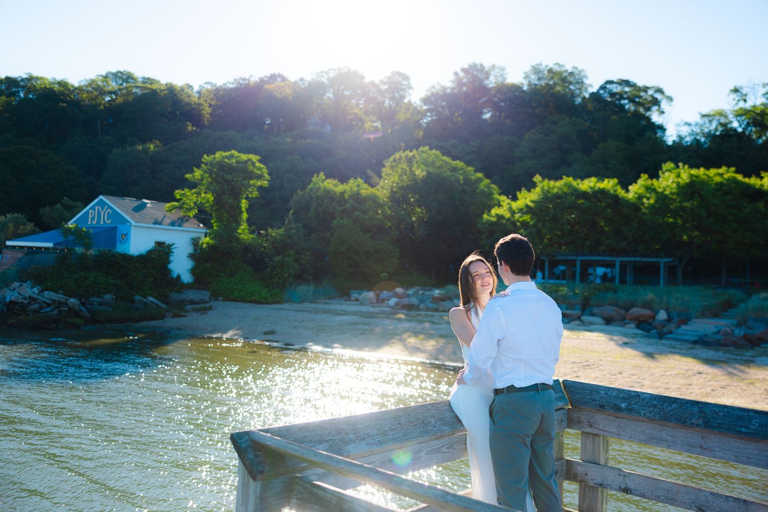 Couple in white standing on a wooden pier overlooking a sunlit cove with a blue cottage and green trees.