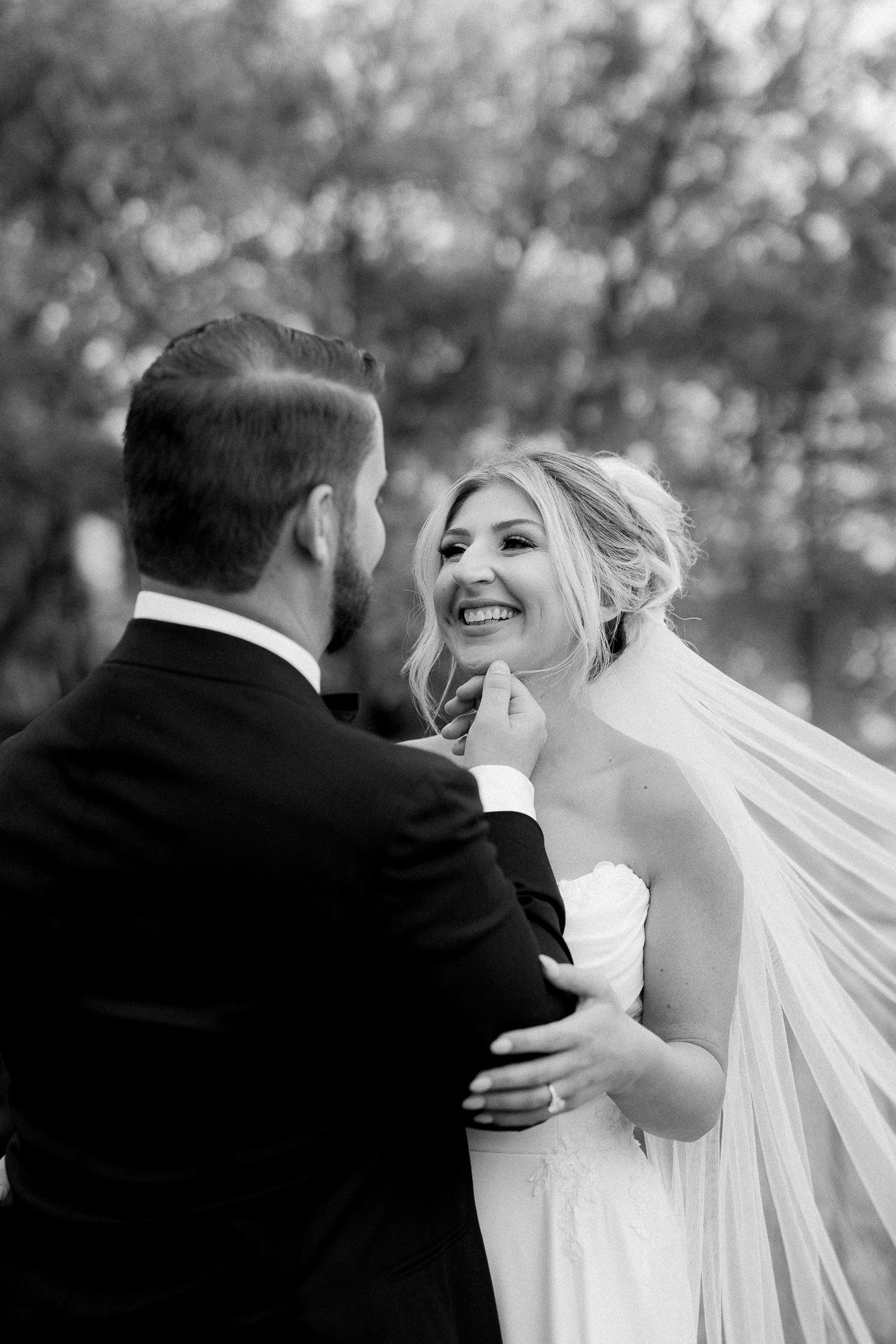 Black and white photo of a smiling bride with flowing veil looking up at her groom.