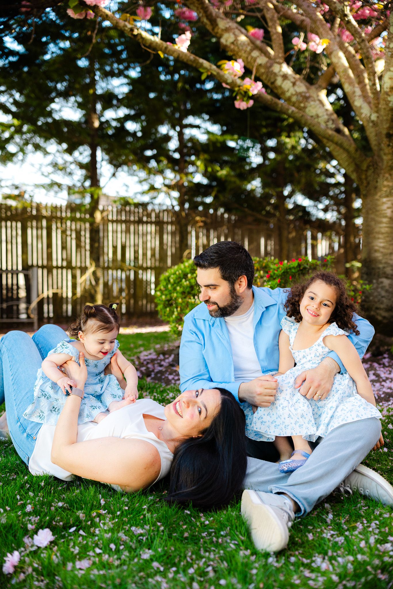 Family of four relaxing on grass under blooming cherry tree, parents and two young daughters in matching blue outfits.