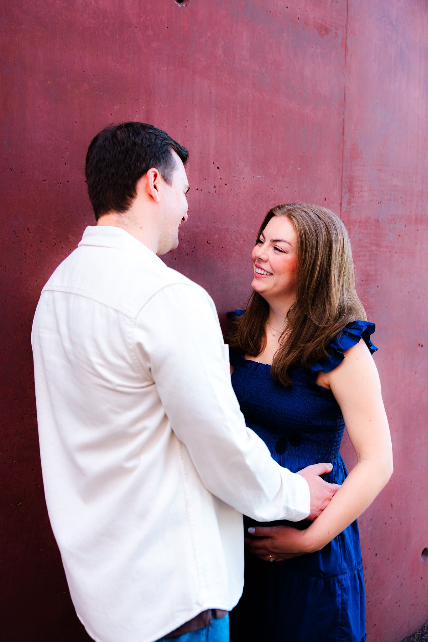 A smiling woman in a blue dress holds hands with a man in a white jacket against a red wall.