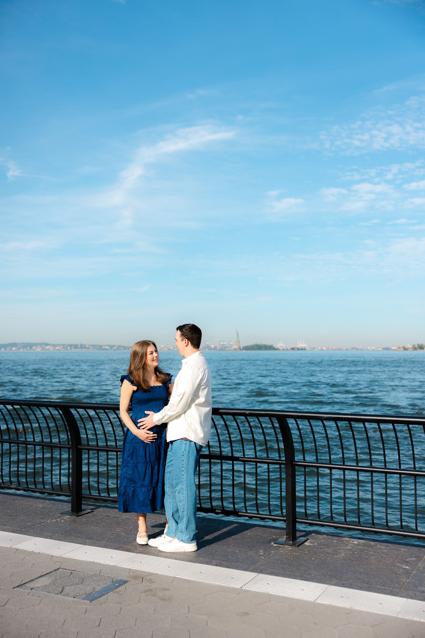 Pregnant couple embracing by waterfront railing with blue sky and harbor views.