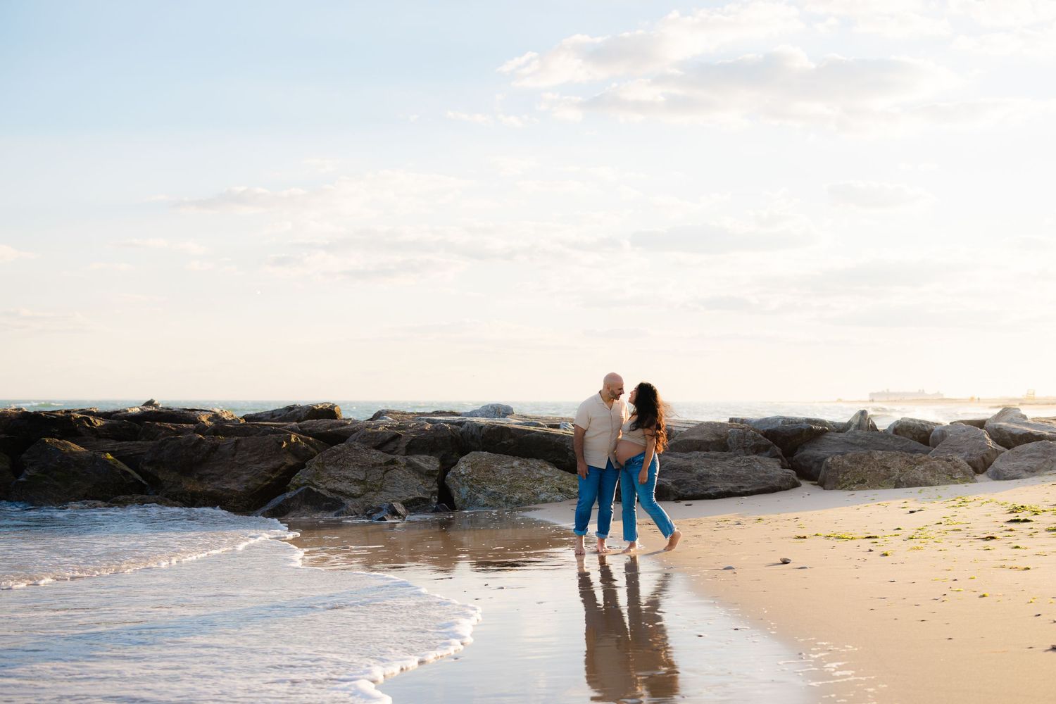Happy couple walking hand-in-hand along a sandy beach with rocky jetty and calm ocean waves at sunset.