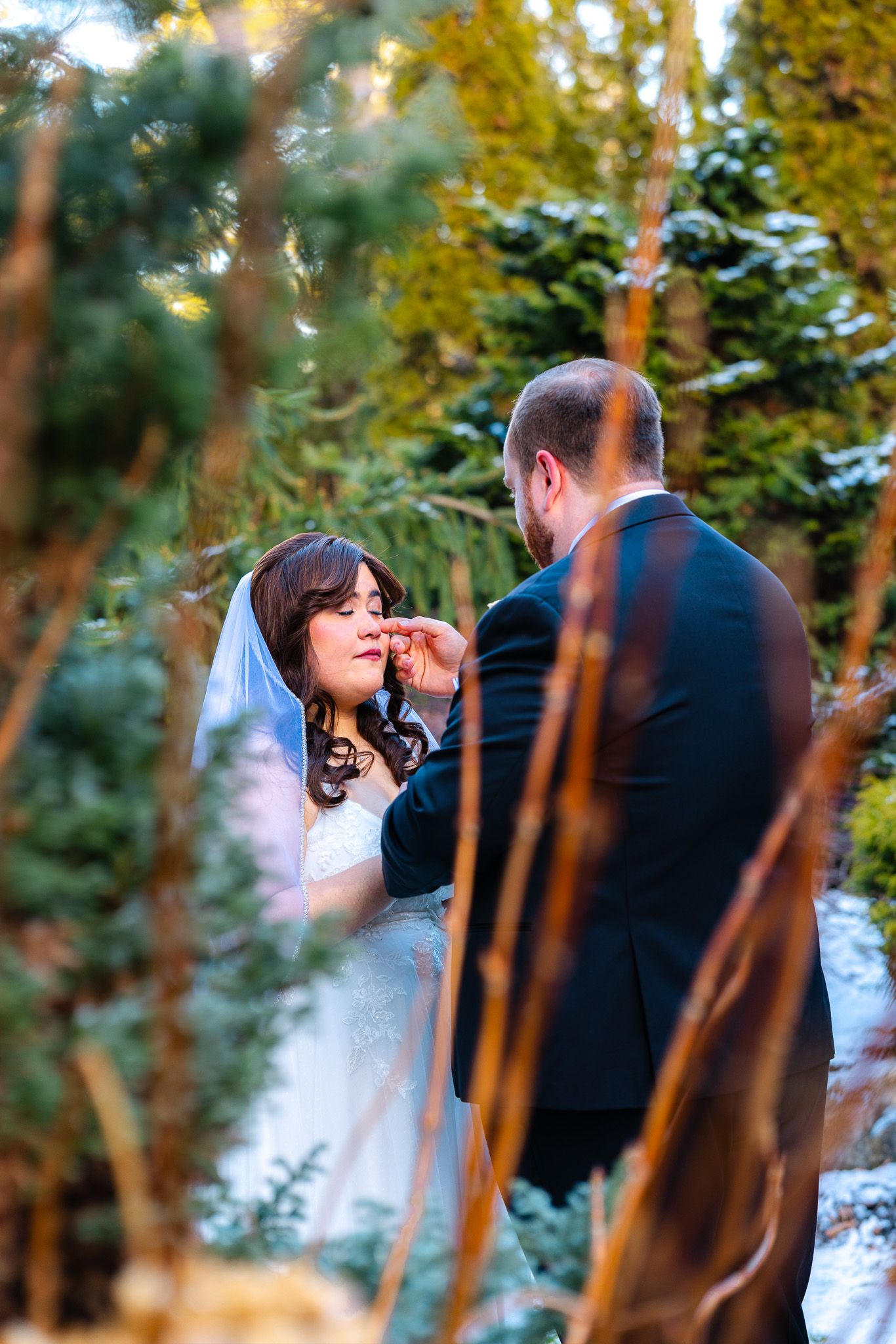 Bride and groom share a tender moment outdoors in a winter garden, surrounded by evergreen trees and snow.